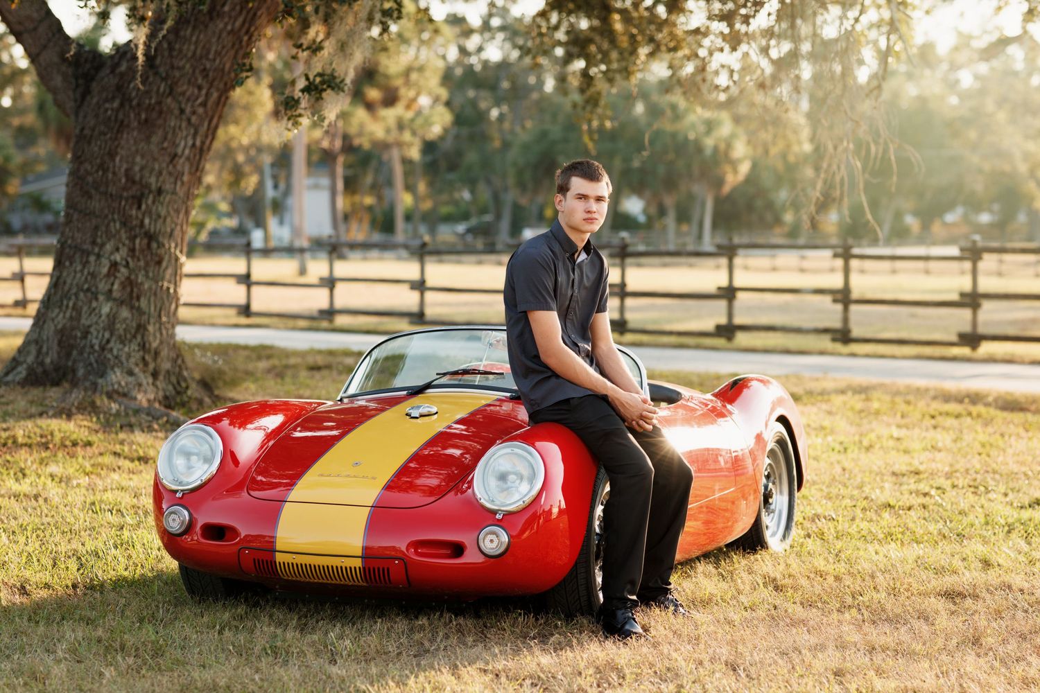 Young man in black polo leaning on a vintage red and yellow Porsche 550 Spyder in a sunny park setting.