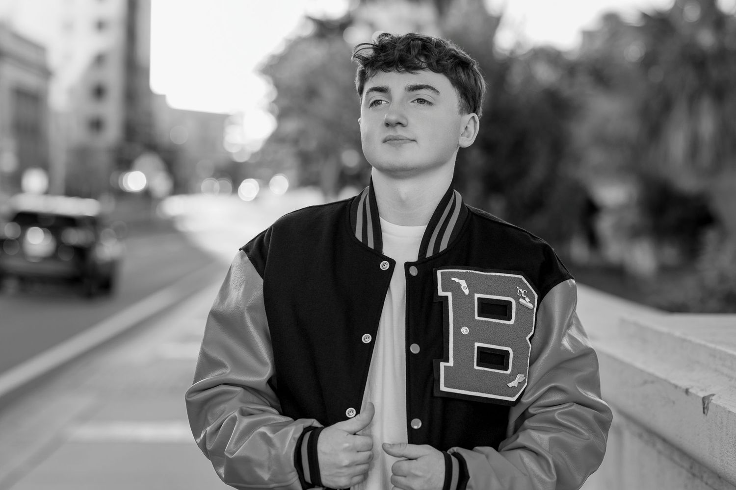 Young man in black and gold varsity jacket posing outdoors in black and white photo.