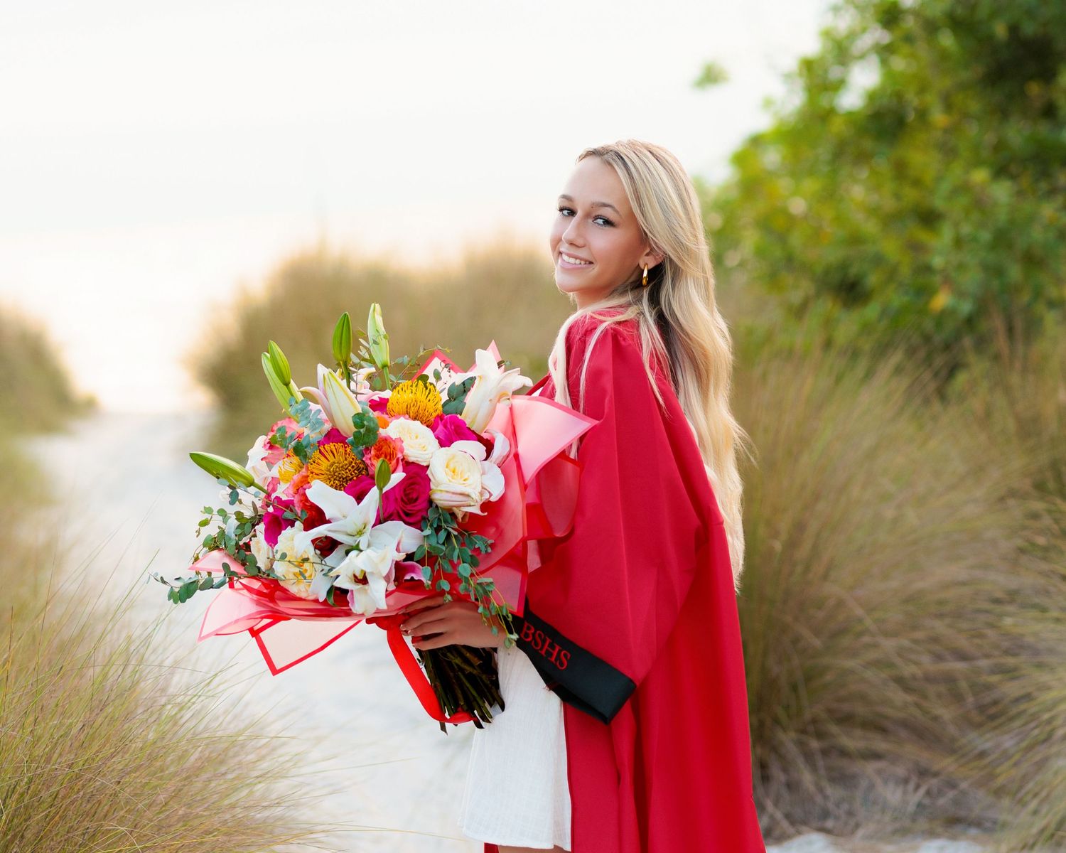Smiling blonde woman in red graduation gown holding colorful bouquet on sandy beach path.