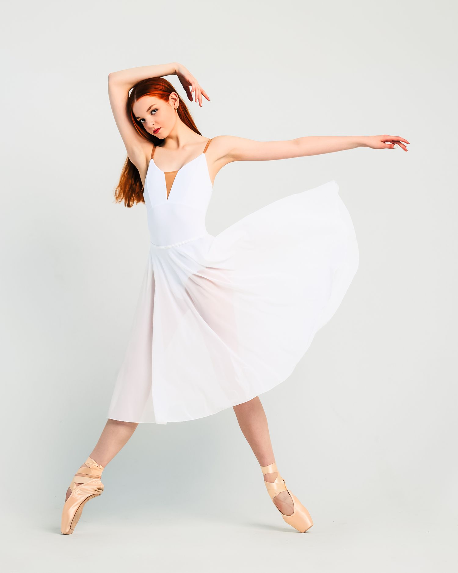 Elegant ballerina in white dress and pointe shoes poses gracefully against light background.