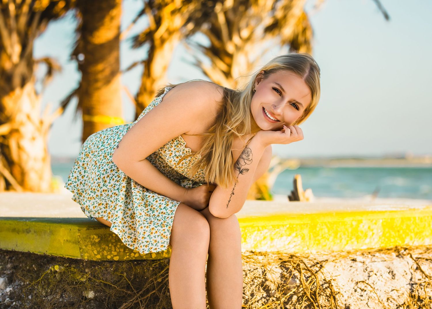 Smiling blonde girl in a floral sundress leans on a colorful wall near a sunny beachside palm setting.