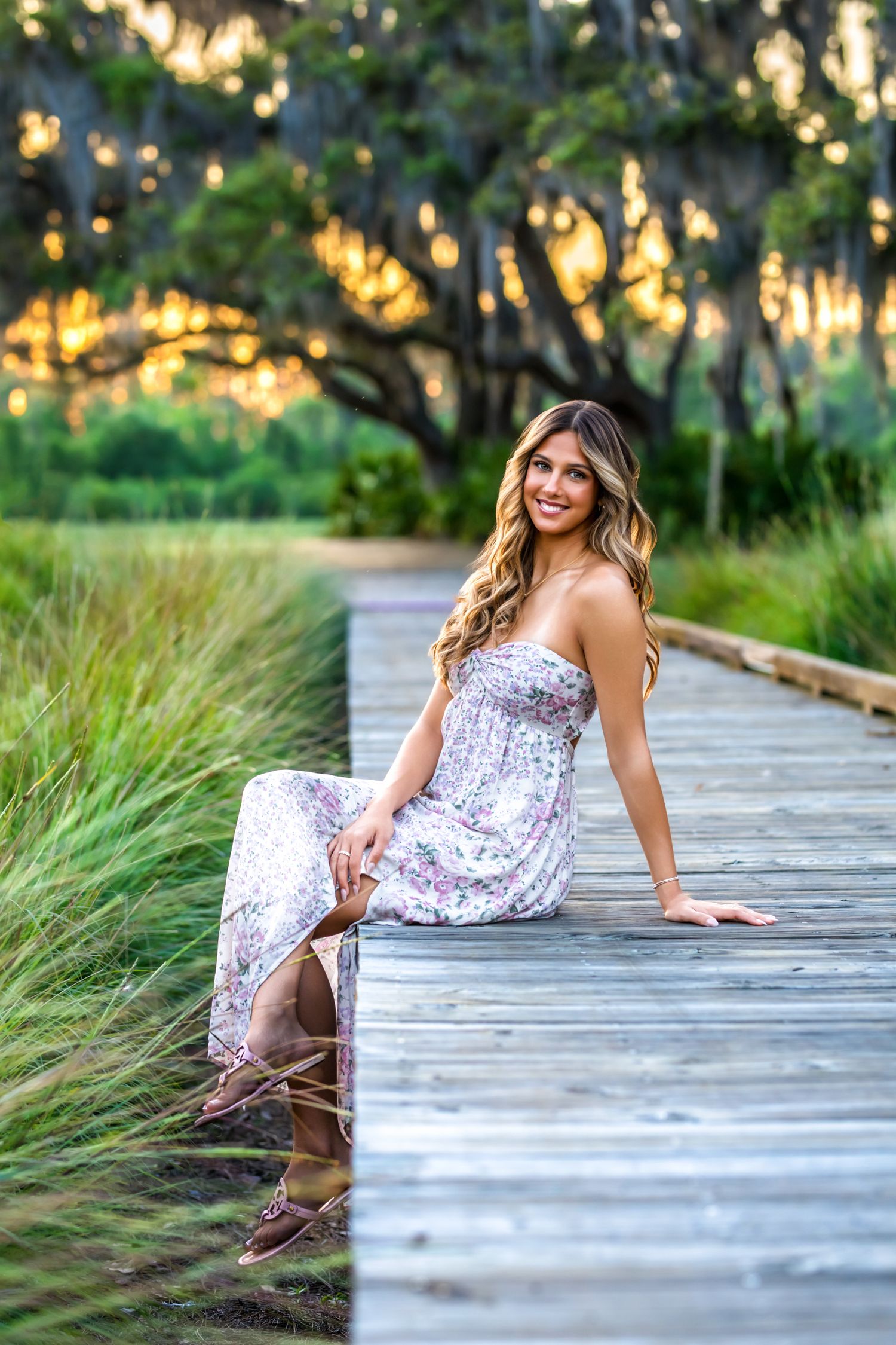 Smiling young woman in a floral strapless dress sits on a wooden boardwalk surrounded by lush greenery.