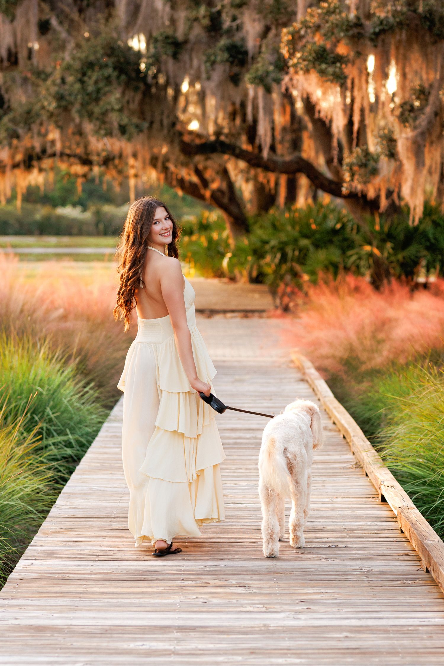 Smiling young woman in a cream tiered gown walks a white dog on a scenic wooden boardwalk at sunset.