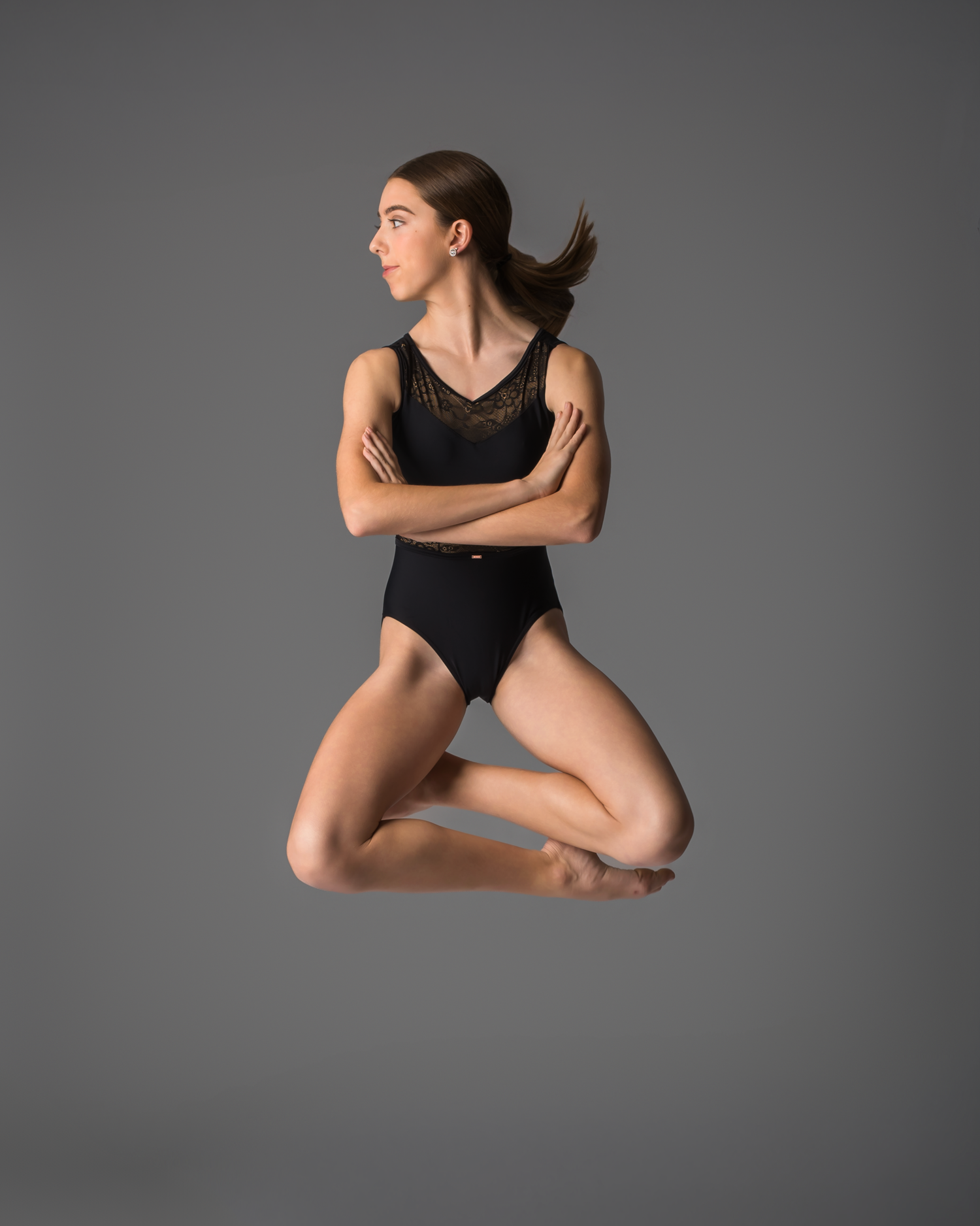 Female dancer jumps with crossed arms in black leotard against gray studio background.