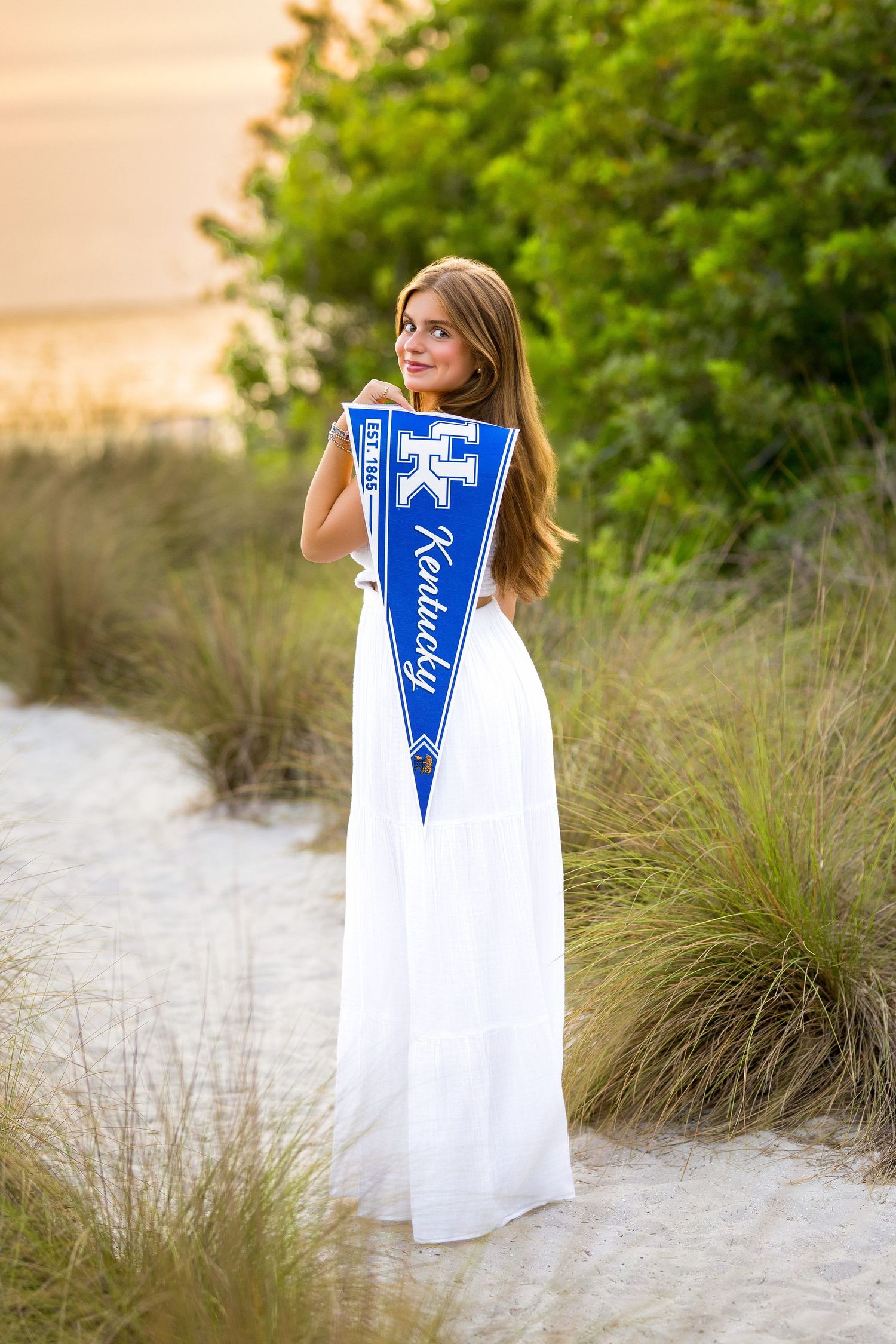 Young woman in white maxi dress holding University of Kentucky pennant on sandy dunes.