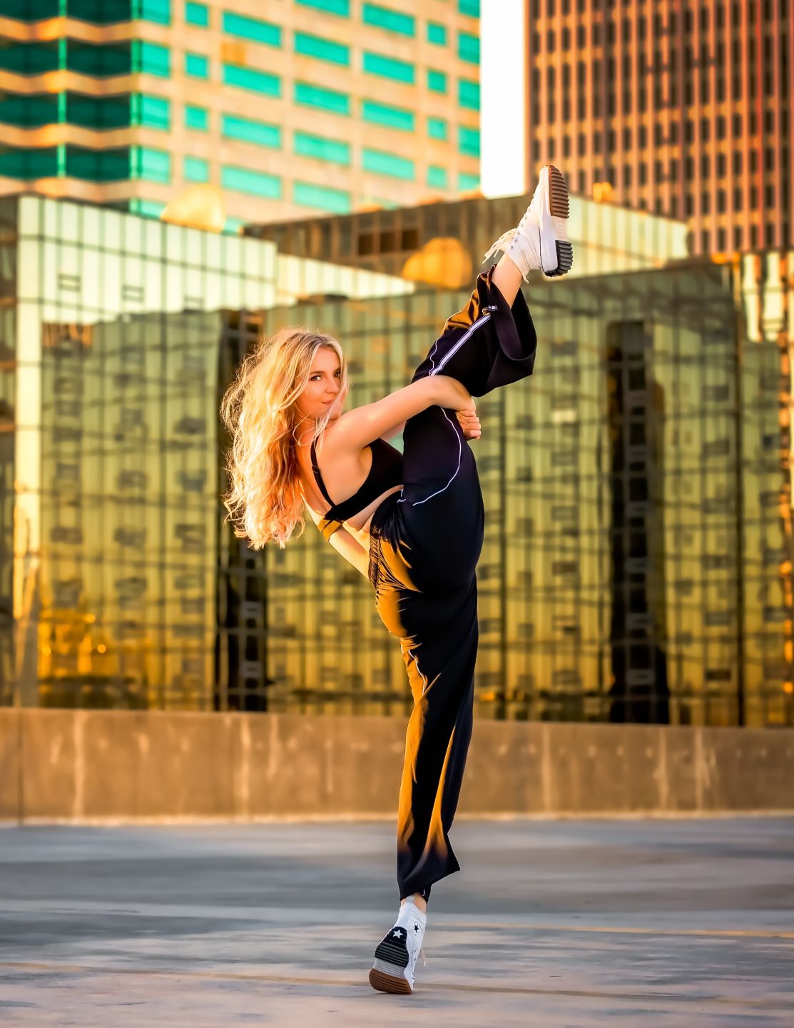 Blonde woman performing a high kick in black athletic wear against golden glass skyscrapers at sunset.