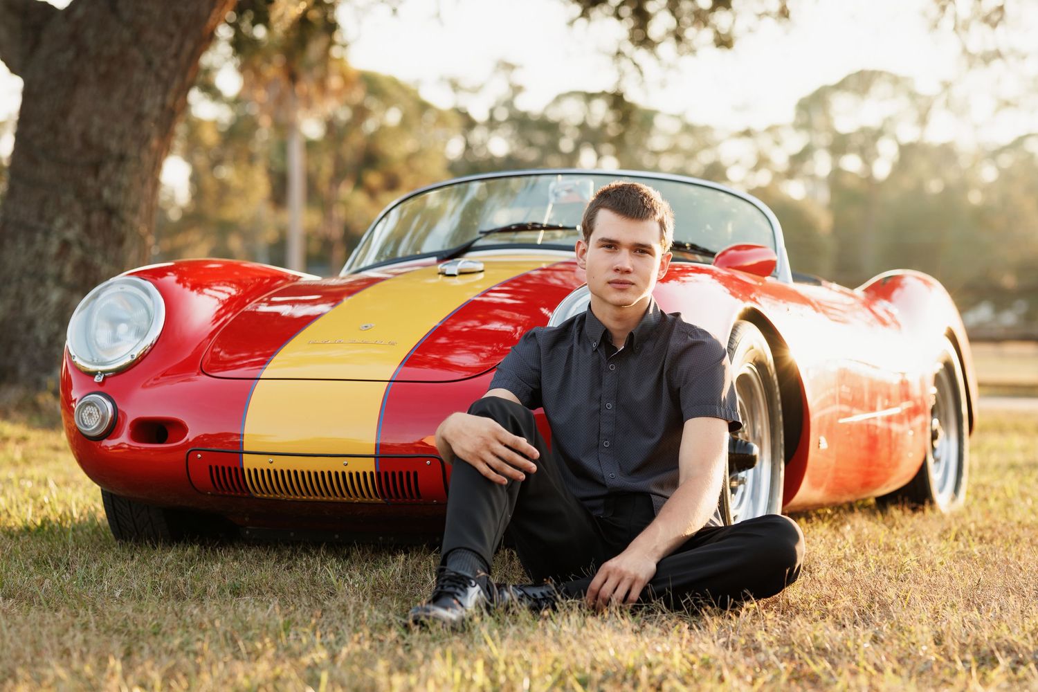 Young man in dark shirt sitting beside a classic red and yellow Porsche sports car outdoors.