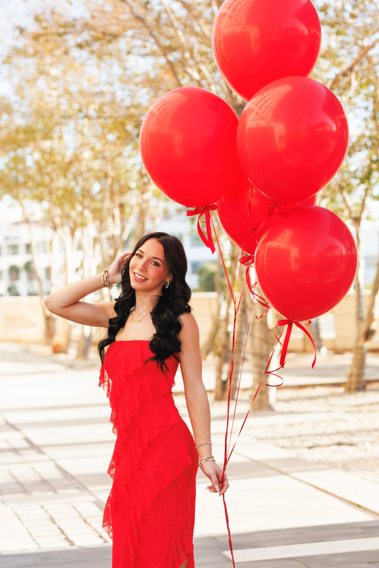 Young woman in red ruffled dress holds large red balloons outdoors on sunny day.