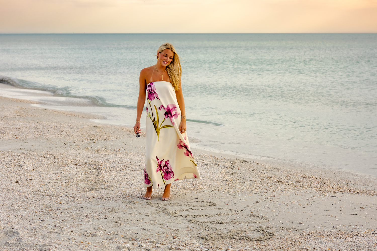 Blonde woman in floral maxi dress walking on sandy beach at sunset.