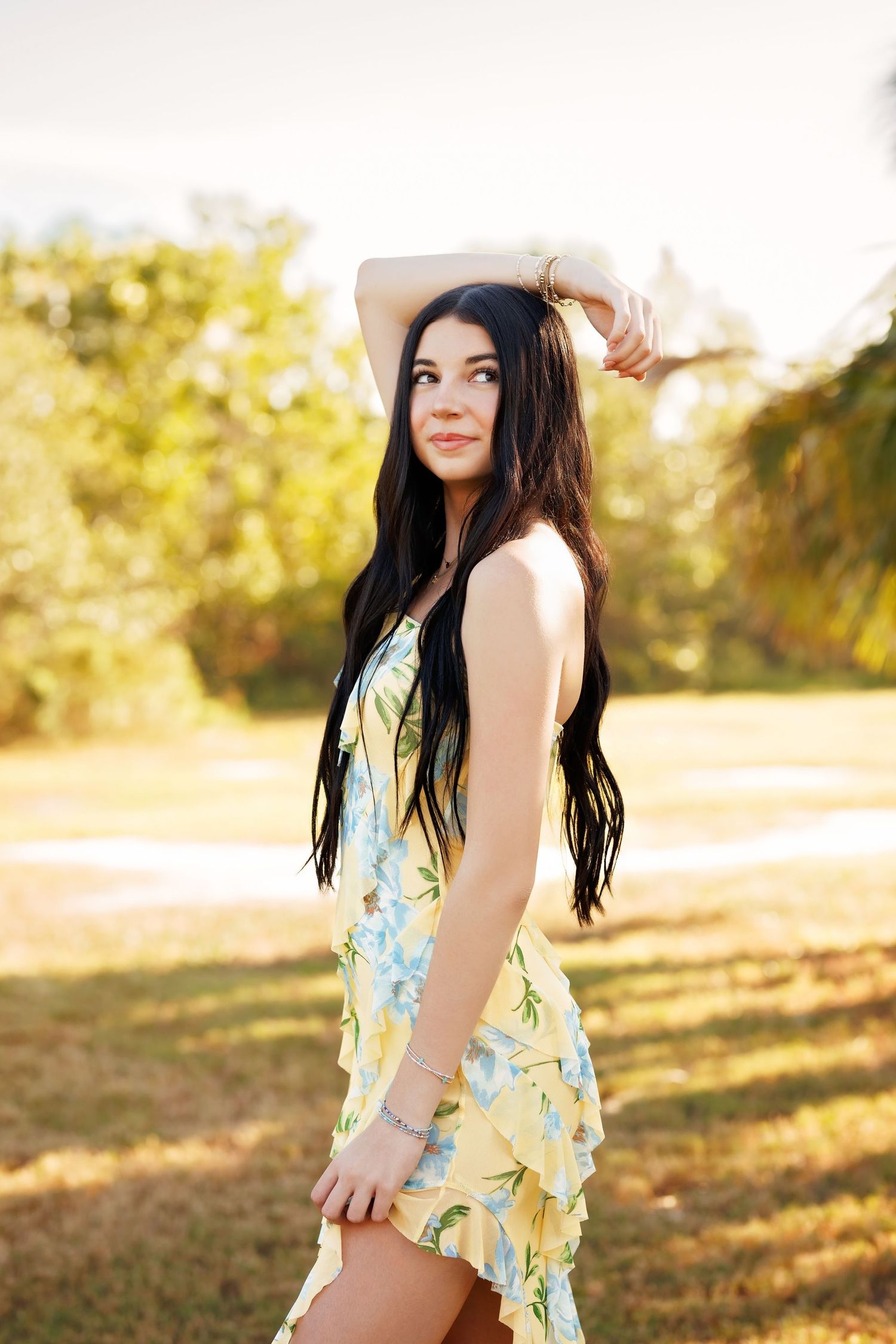 Young woman in yellow floral sundress posing outdoors in a sunlit park with long dark hair.