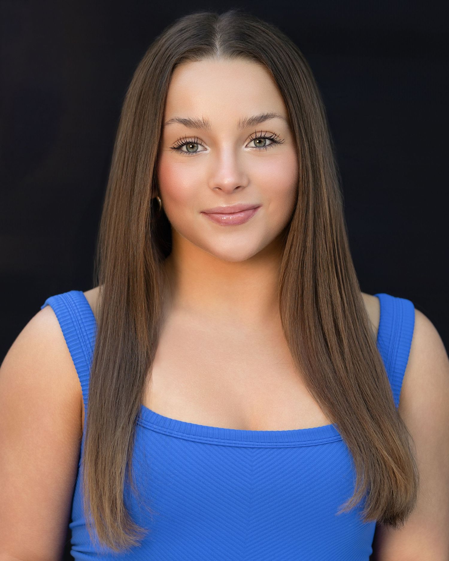 Young woman with long brown hair wearing a blue top poses for a professional headshot photo against dark background.