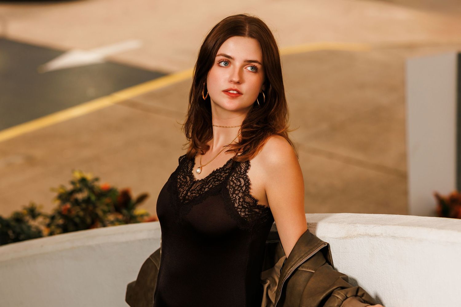Young woman in black lace camisole and olive jacket posing outdoors in urban setting.