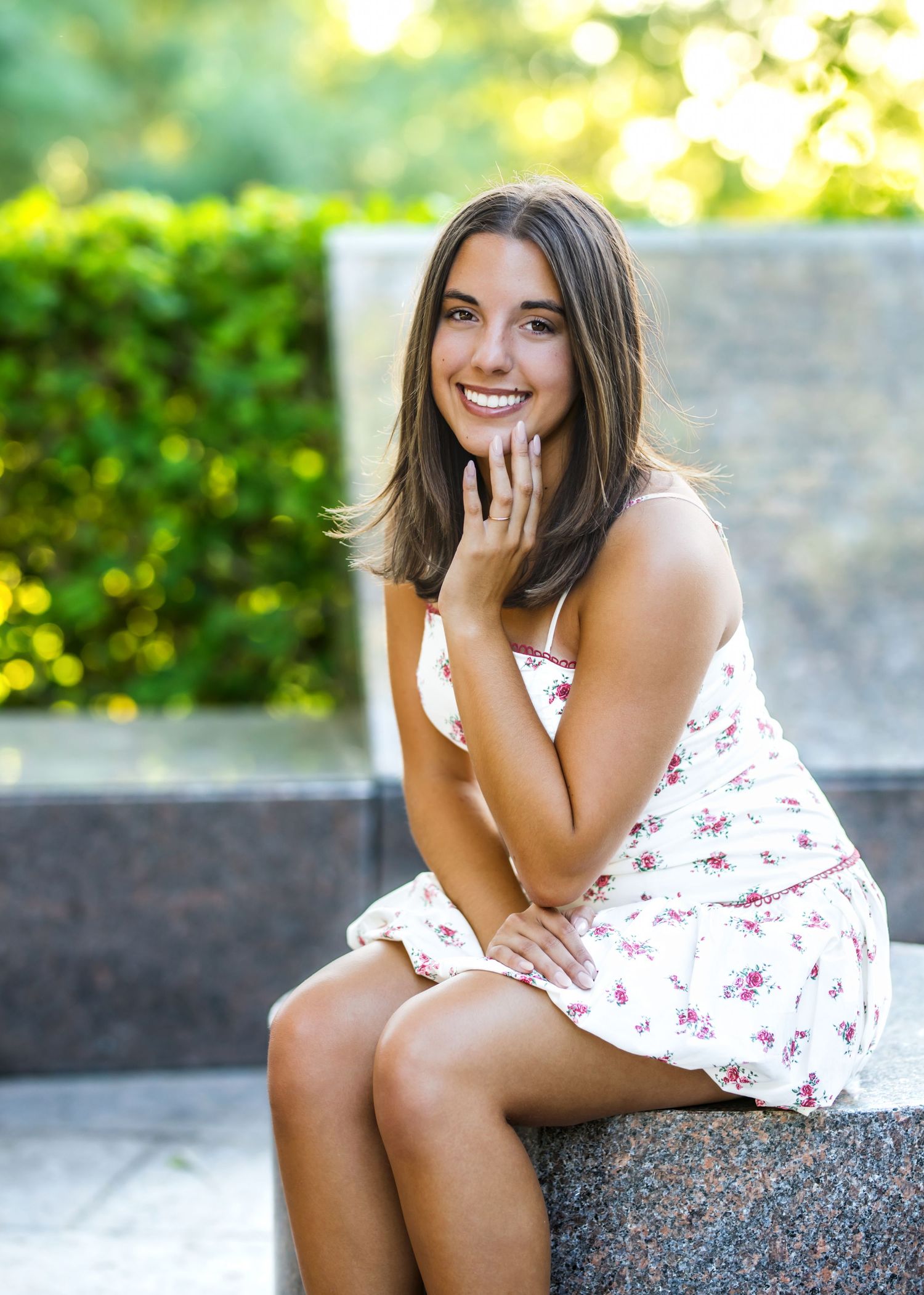 Smiling teenage girl in floral sundress sitting outdoors in lush green garden setting.