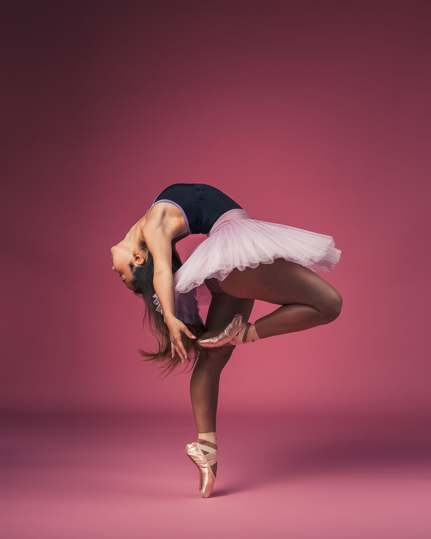 Ballerina balances on pointe in pink tutu with dramatic backbend against rose-colored background.