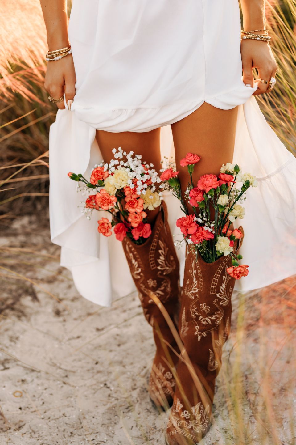 Woman in white dress and brown cowboy boots with floral arrangements tucked inside, standing on sandy path.