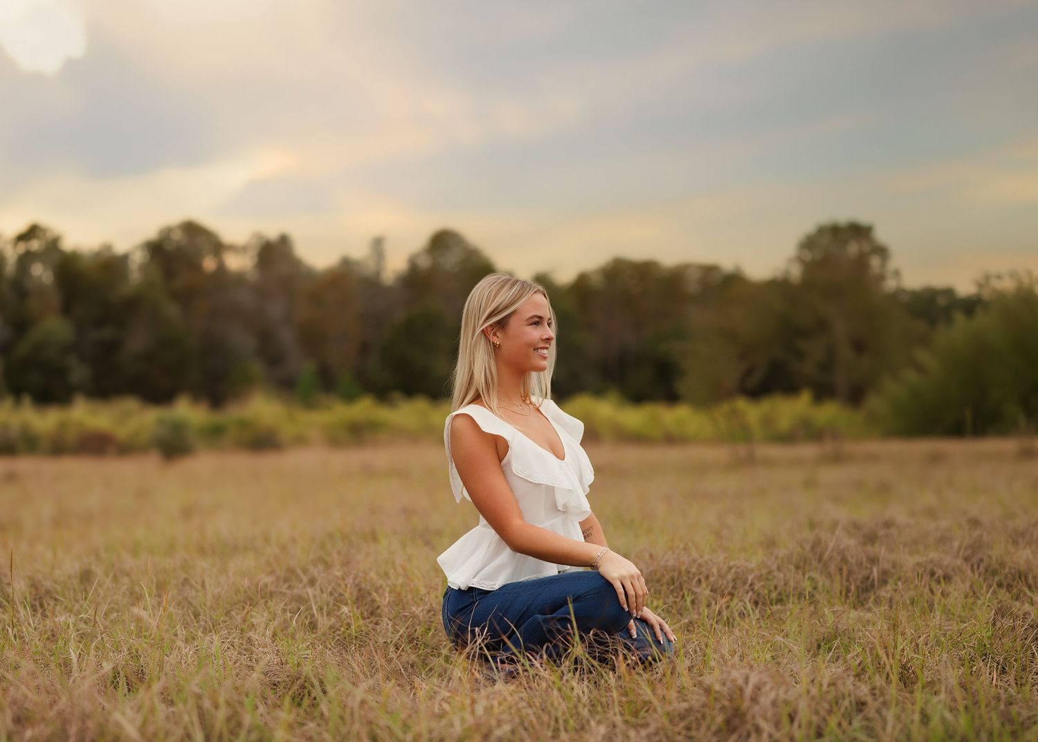 A blonde high school senior girl in a white top and jeans sits peacefully in a field at golden hour with trees in the background in Tampa.