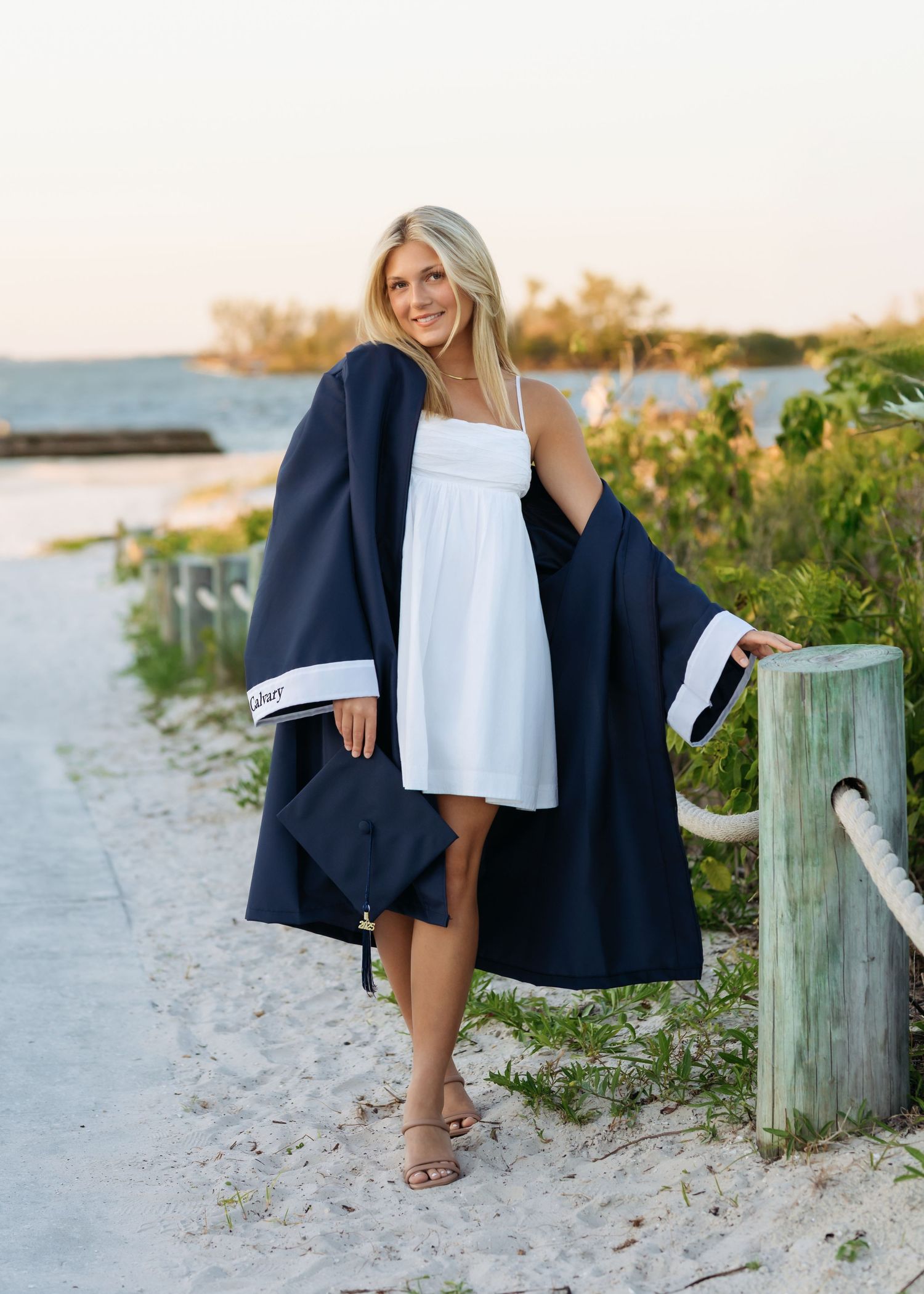 Blonde woman wearing navy graduation gown over white dress on sandy beach pathway at sunset.