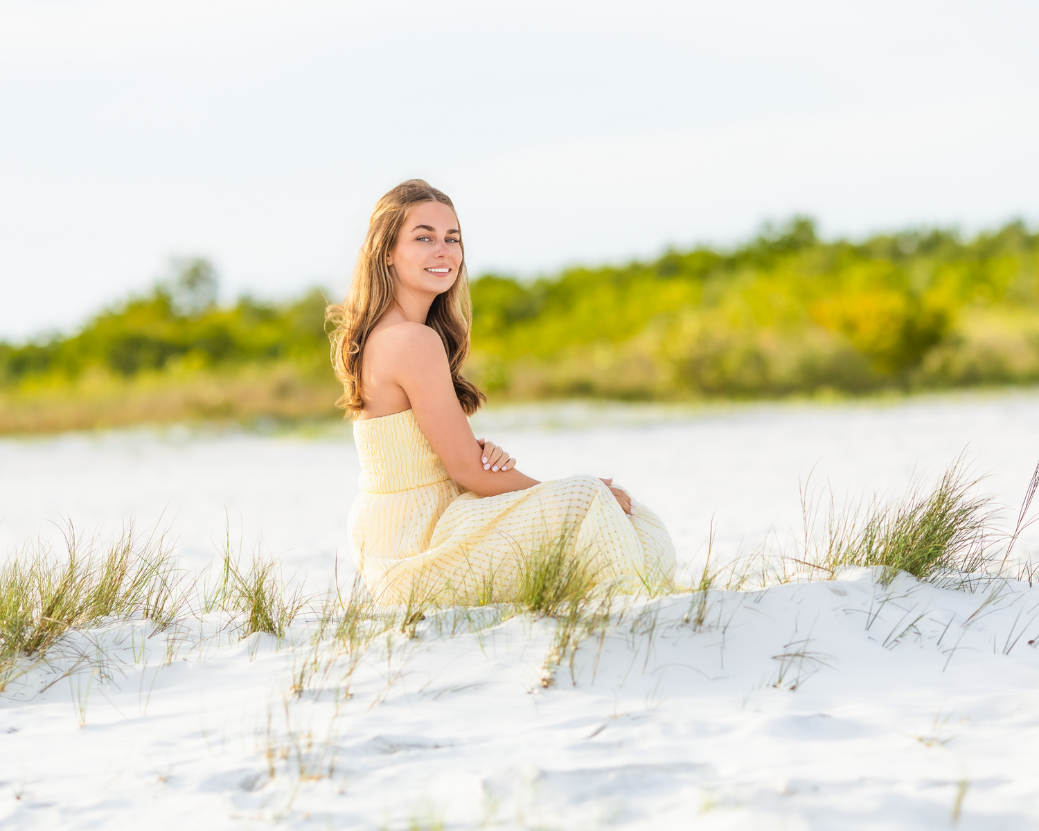 Smiling brunette woman in yellow sundress sitting on white sand dunes near coastal greenery.