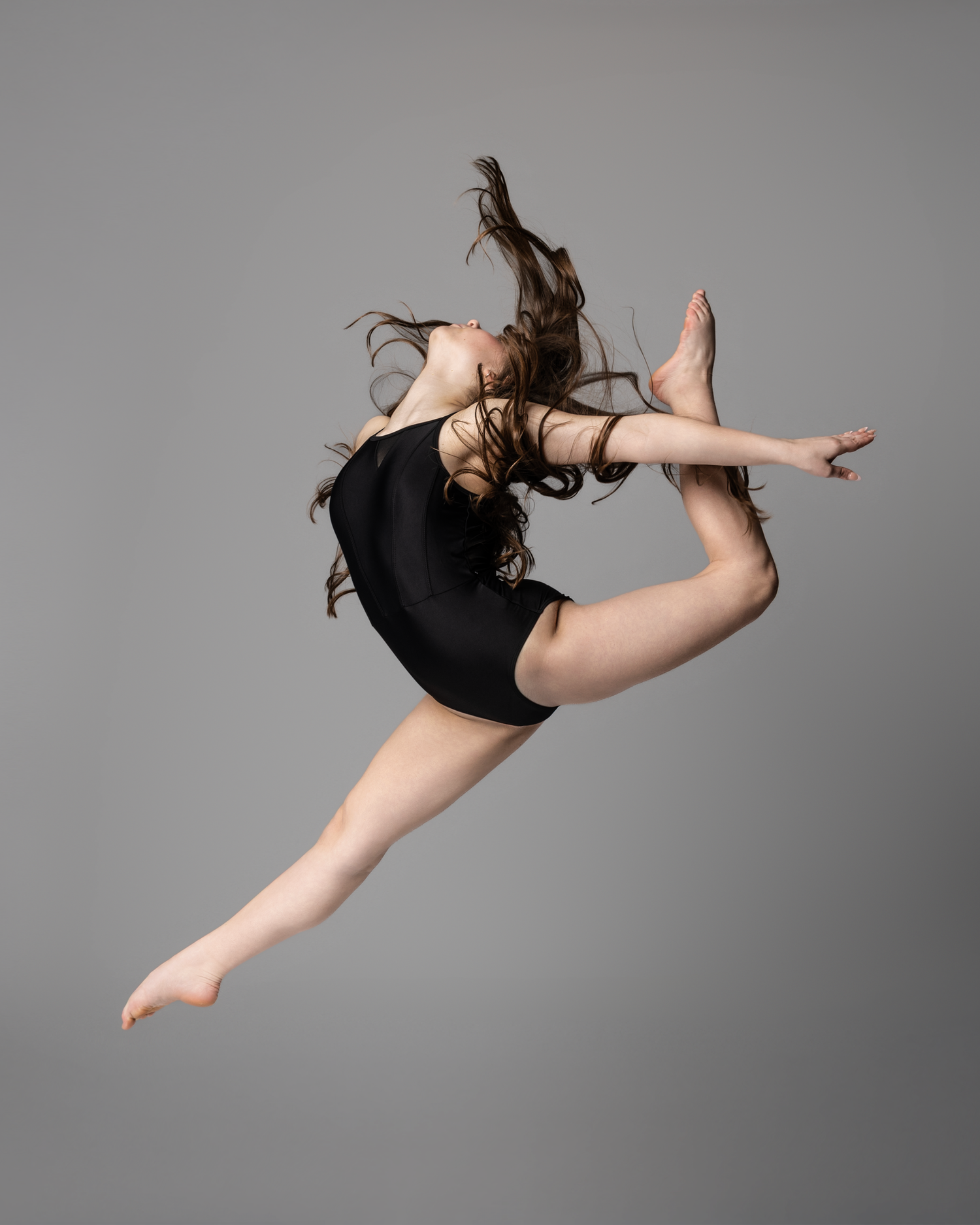 Dancer leaps gracefully in black leotard with hair flowing against gray studio background.