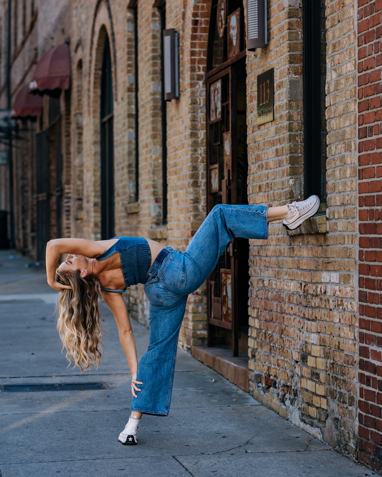 Flexible dancer kicks leg high while leaning against brick wall in wide-leg denim overalls.