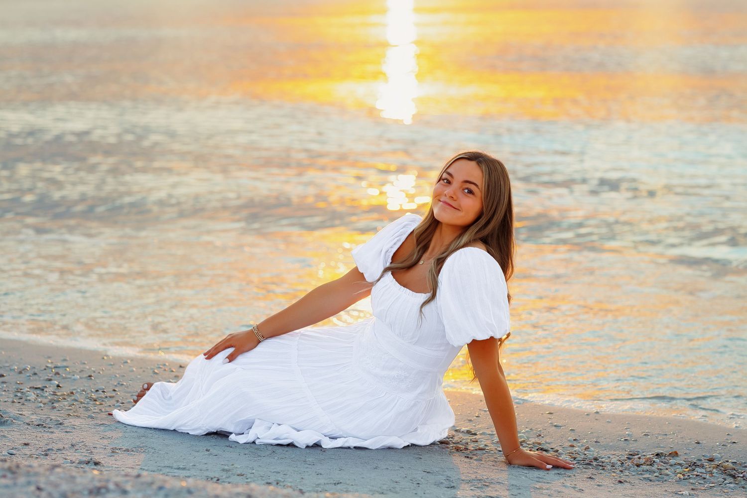 High school senior in white dress sitting on sandy beach in Tampa at golden sunset with ocean waves in background.