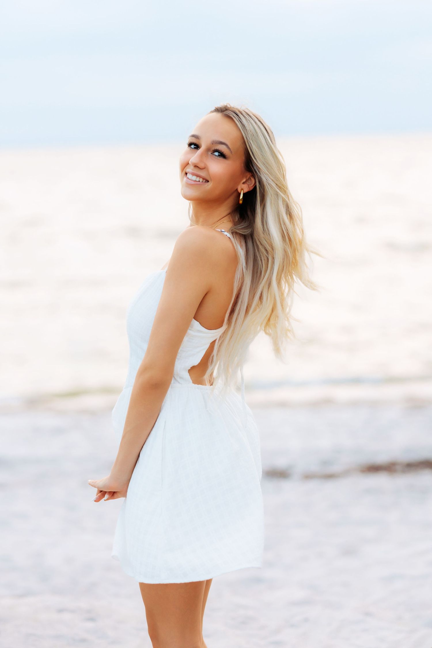 Smiling blonde high school senior in white sundress standing on beach in Tampa with ocean waves in background.