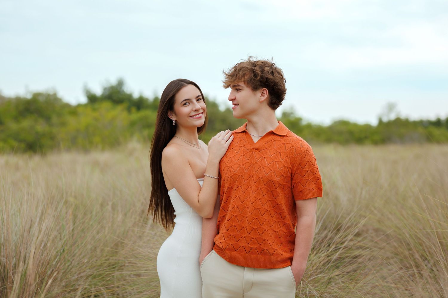 high school senior sweethearts pose romantically in a grassy field, woman in white dress, man in orange polo shirt.
