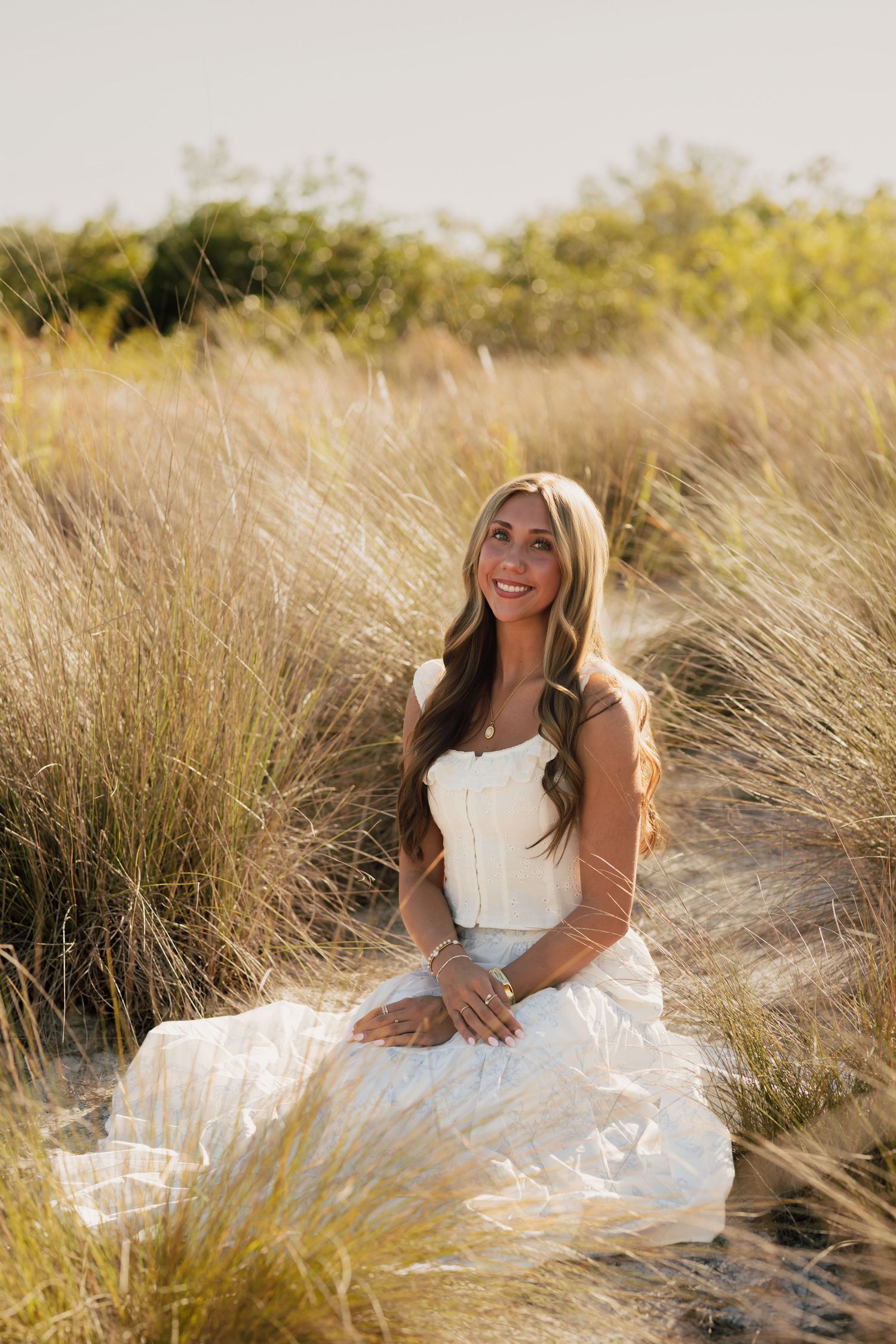 Smiling woman in white dress sitting in golden beach grass dunes at sunset.