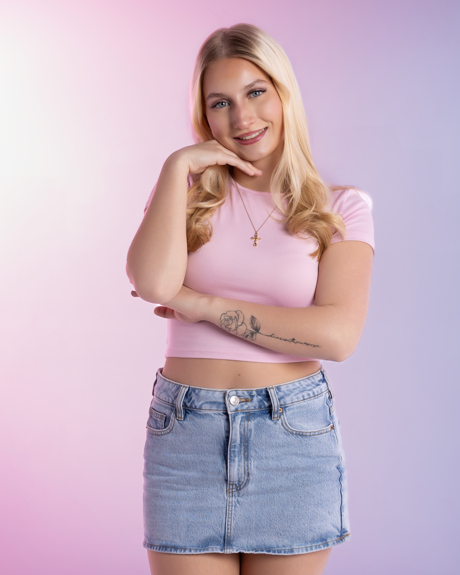 Blonde woman in pink crop top and denim mini skirt posing against purple gradient studio background.
