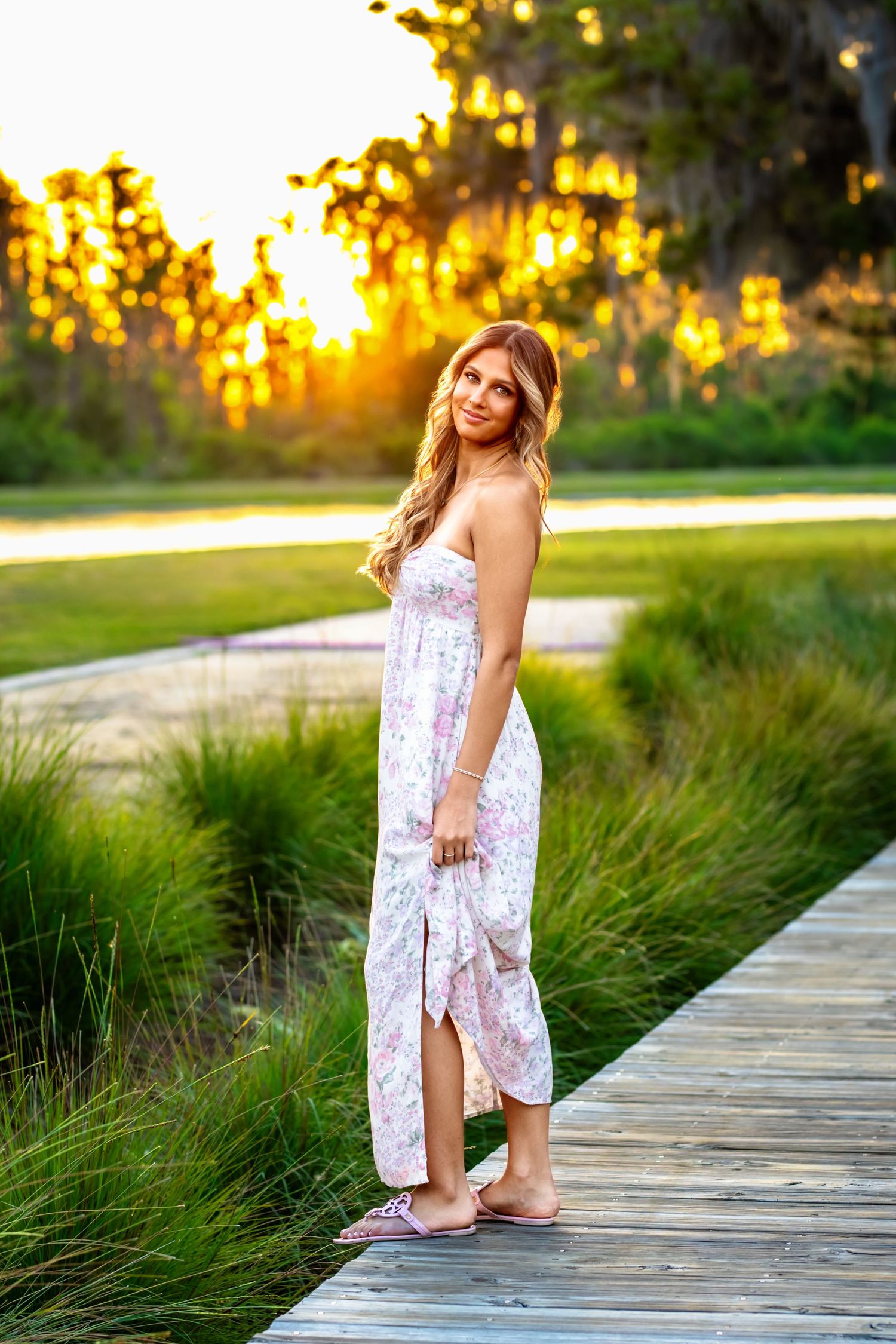 Blonde teenage girl in floral sundress stands on wooden boardwalk at golden sunset.