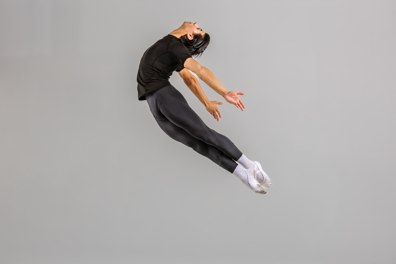 Male dancer leaps dramatically in black outfit against a plain gray studio background.
