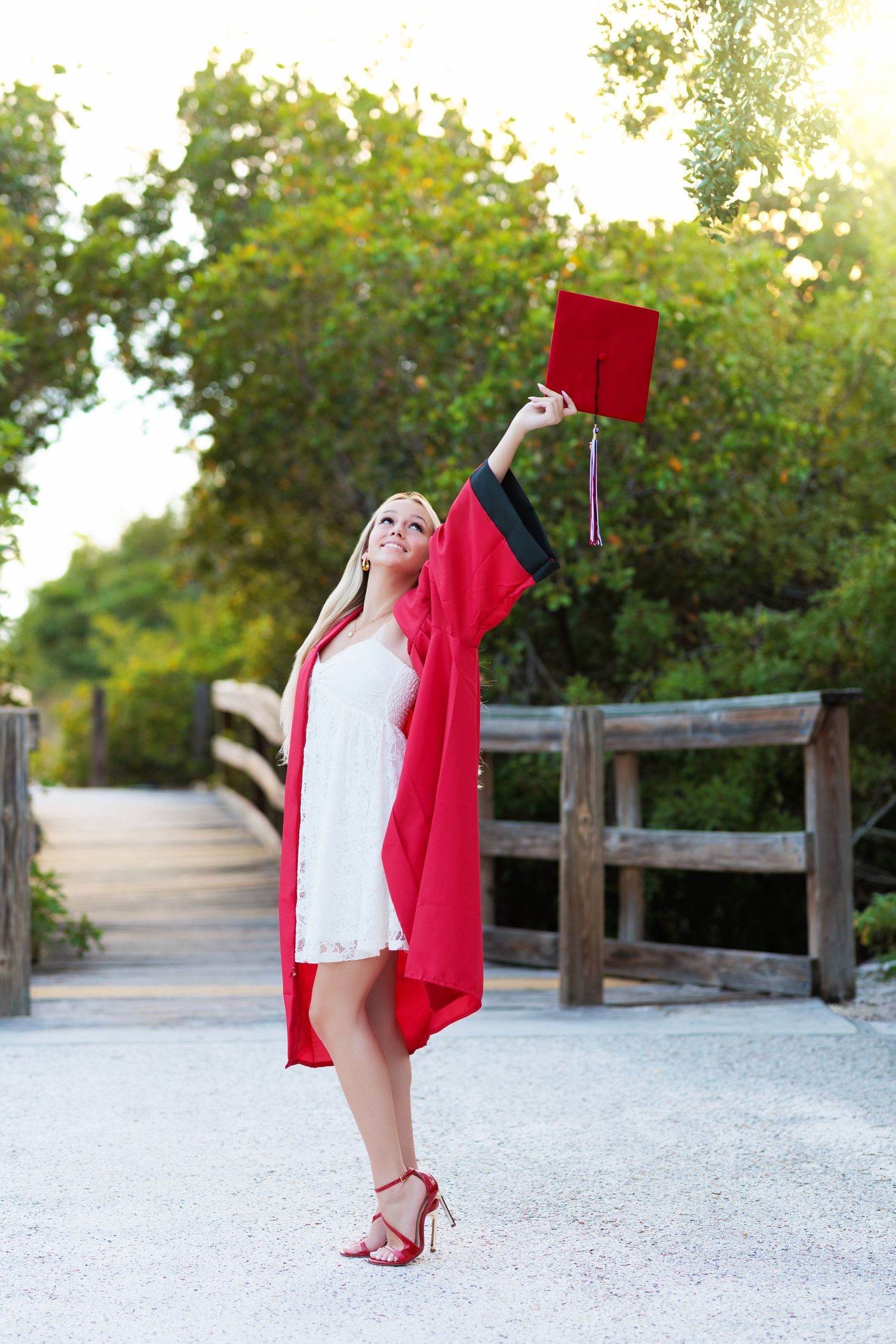 Graduate in red cap and gown tossing cap in air on sunny outdoor walkway.