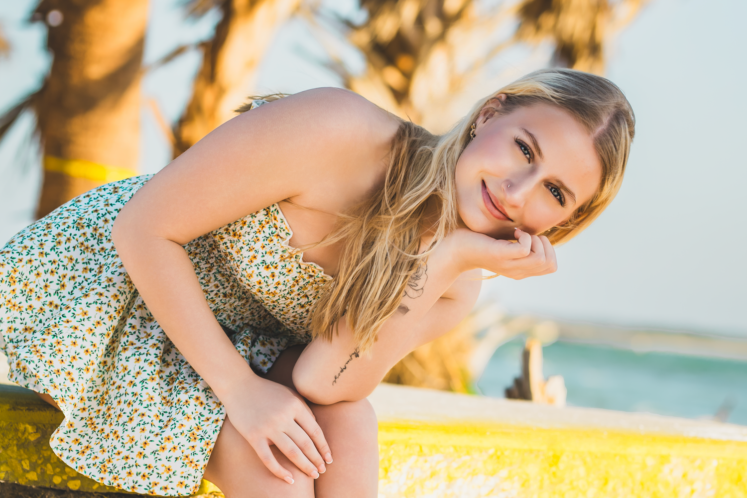 Smiling blonde woman in floral sundress sitting outdoors near colorful yellow wall on sunny day.