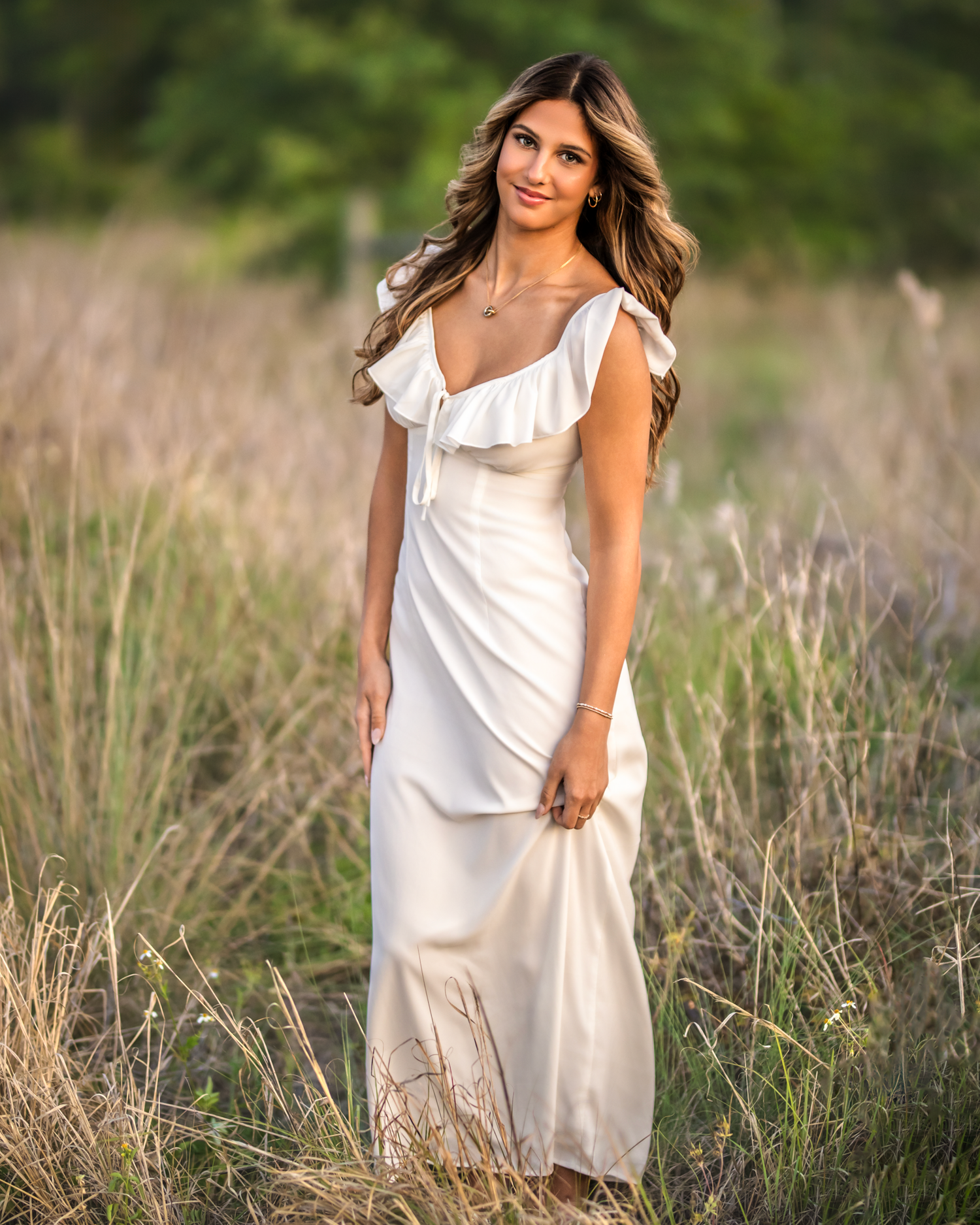 Young woman in elegant white maxi dress with ruffle neckline posing in tall grass field during golden hour.