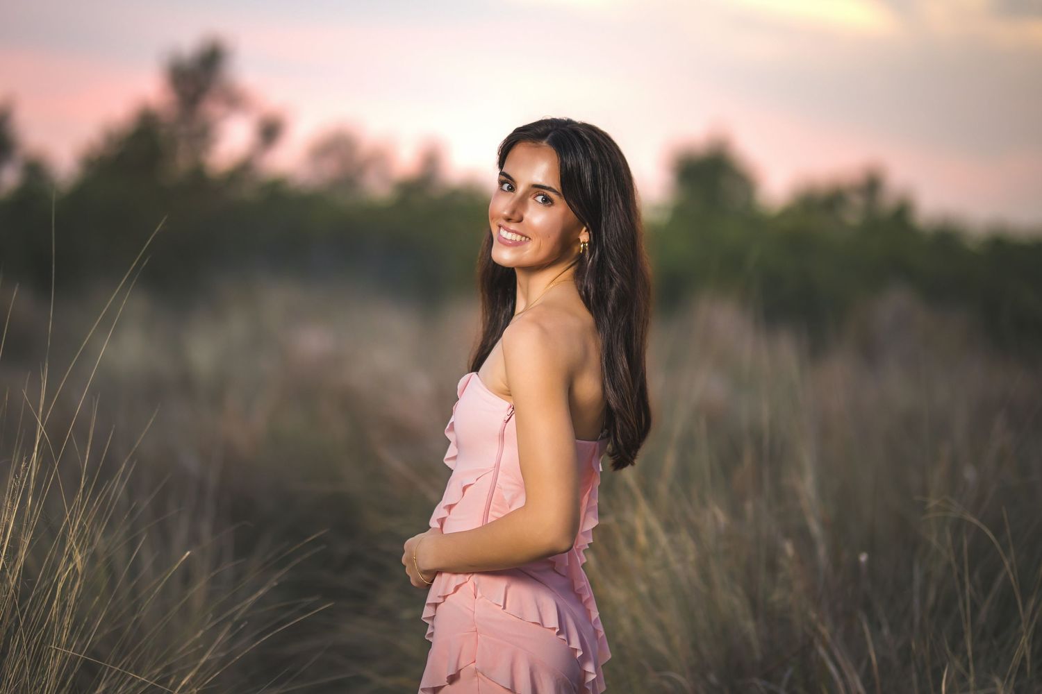 Brunette woman in pink strapless dress smiling over shoulder in tall grass field at sunset.
