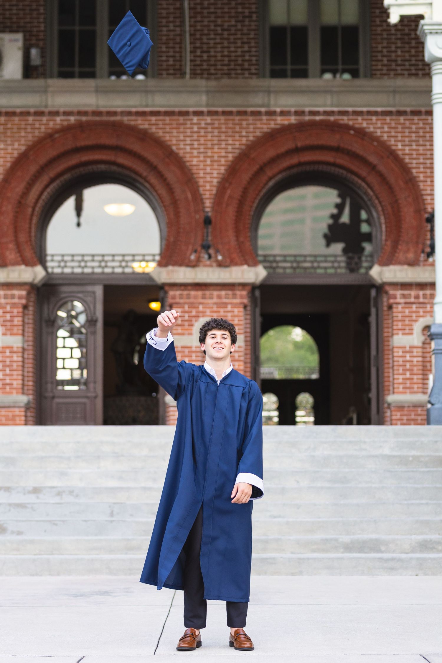 Young man in blue graduation gown tossing cap in front of brick university building steps.