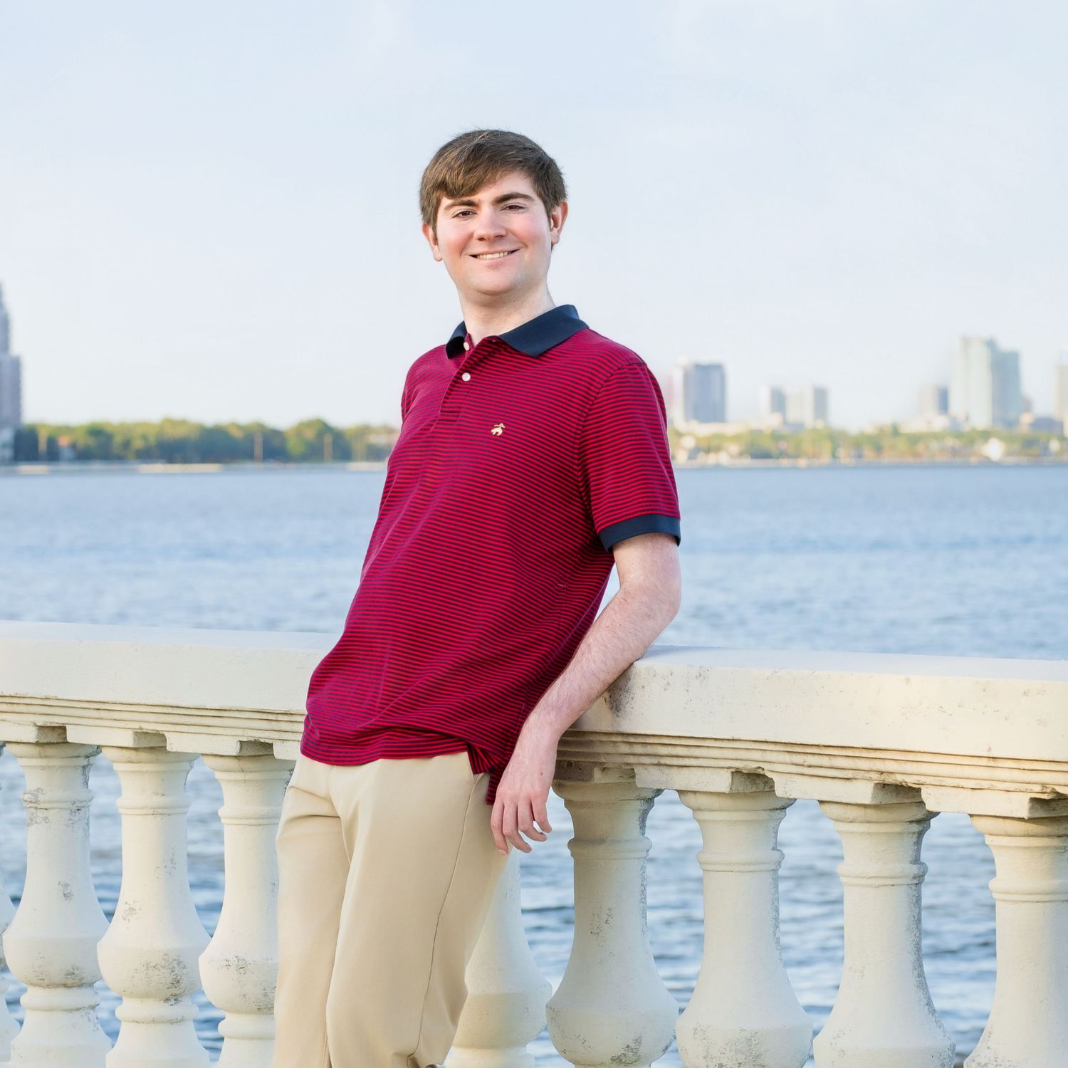 Young man in red polo shirt leaning on white balustrade with city skyline and waterfront in background.