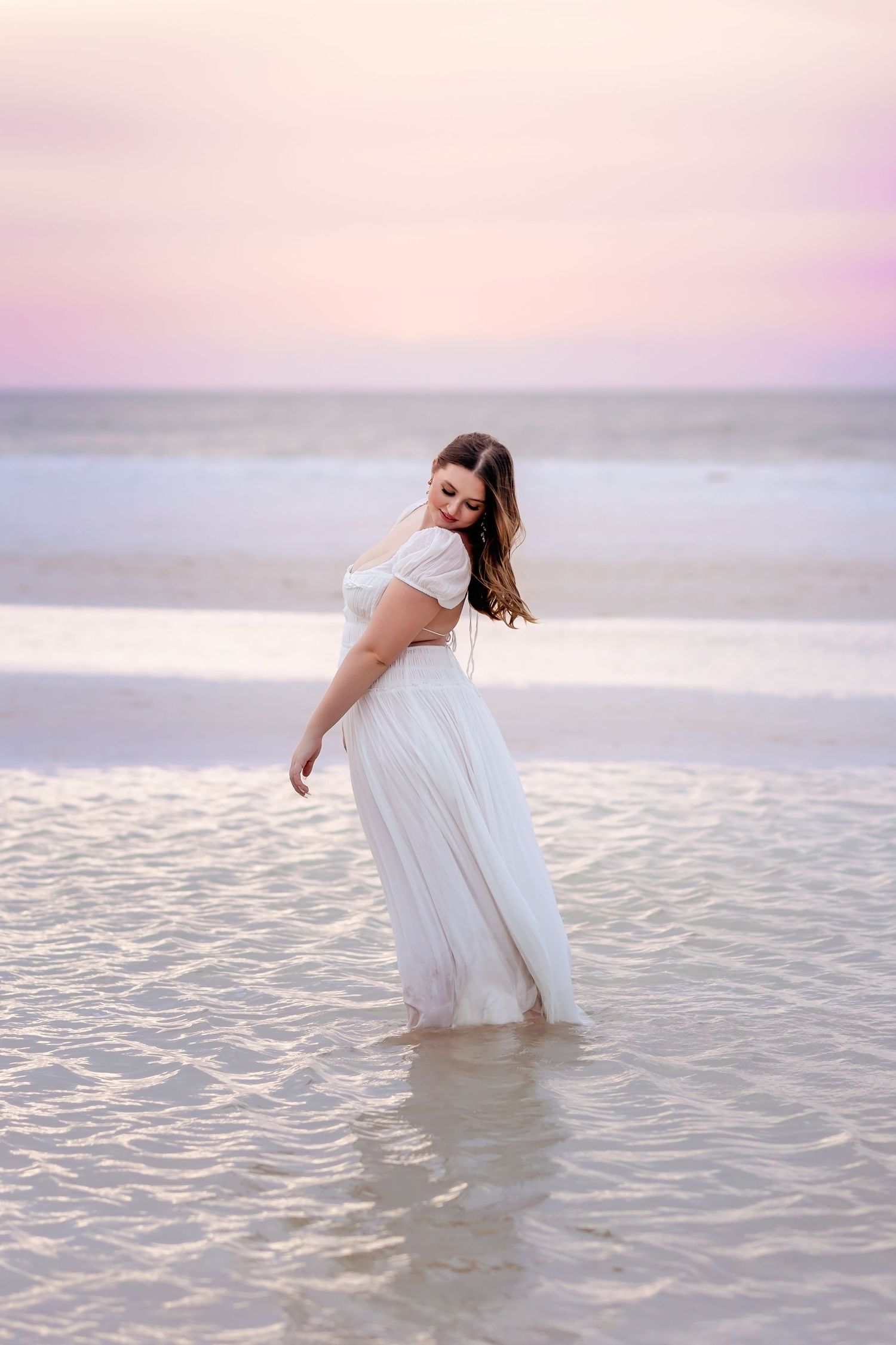 Young woman in white off-shoulder dress standing in shallow ocean water at sunset.