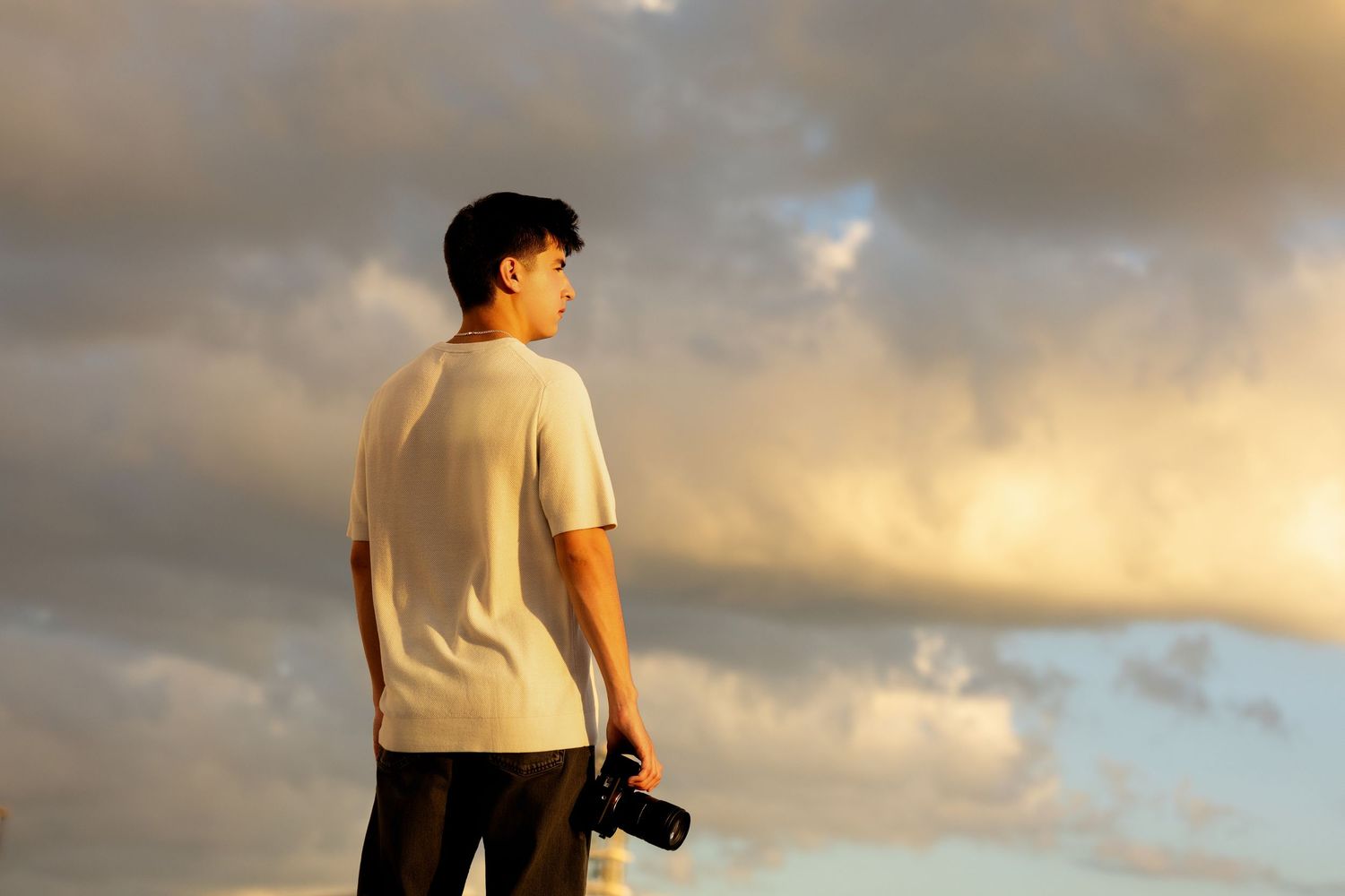 Young man holding camera standing against dramatic golden cloudy sky at sunset.