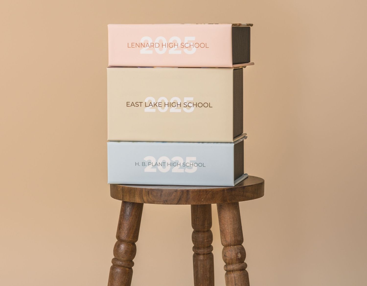 Three stacked high school senior portrait boxes labeled with school names on a wooden stool.