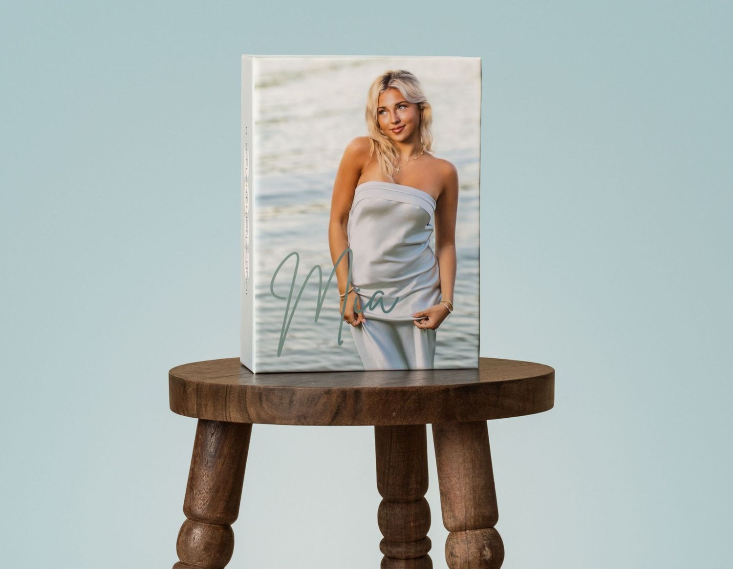 A photo book featuring a smiling blonde woman at the beach displayed on a rustic wooden stool.