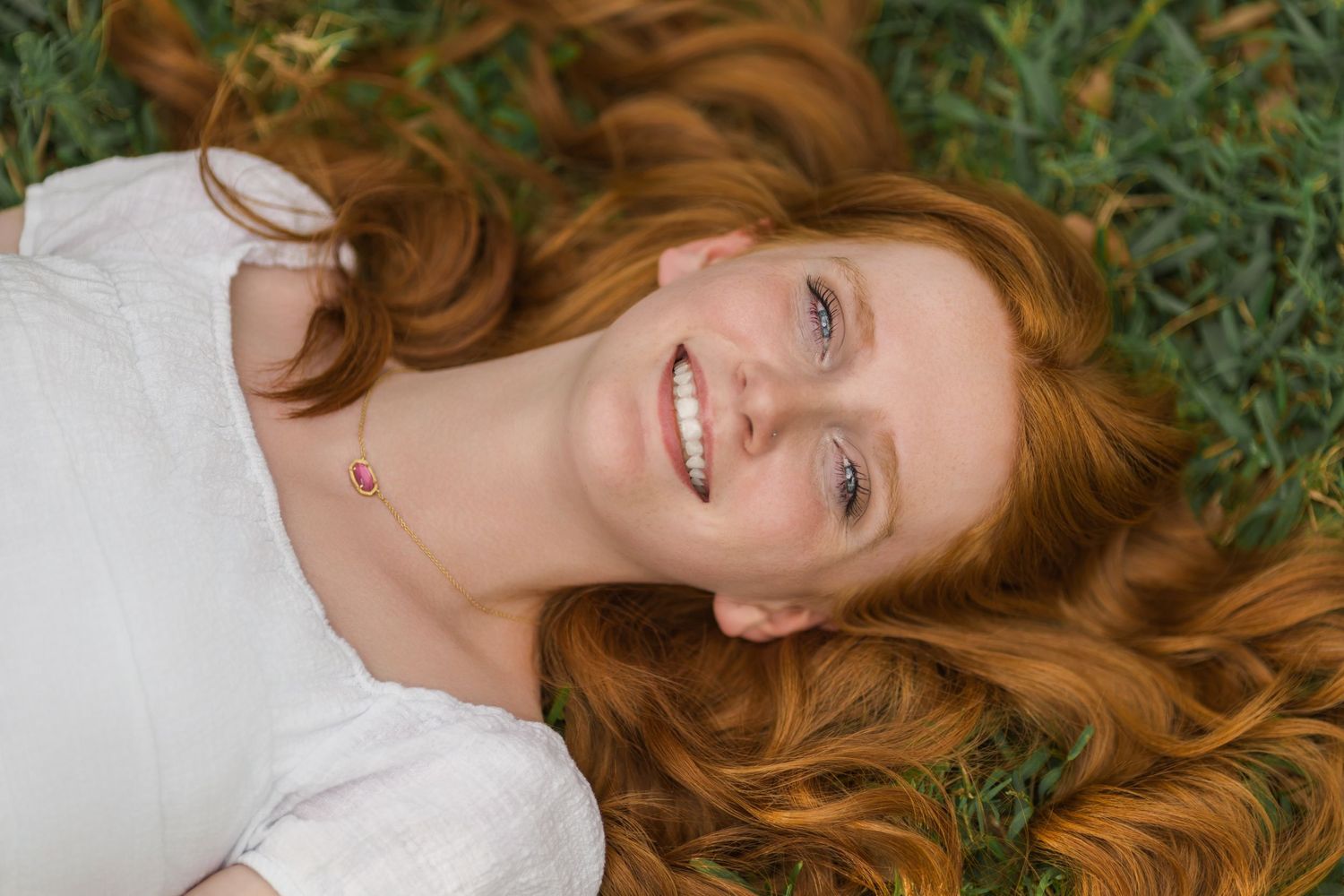 Smiling redhead woman lying on green grass with long wavy hair spread around her.