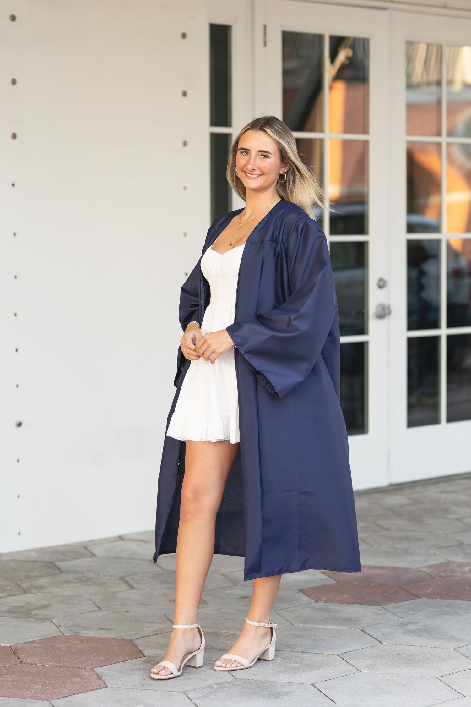 Smiling blonde woman in navy graduation gown and white dress posing outside building entrance.