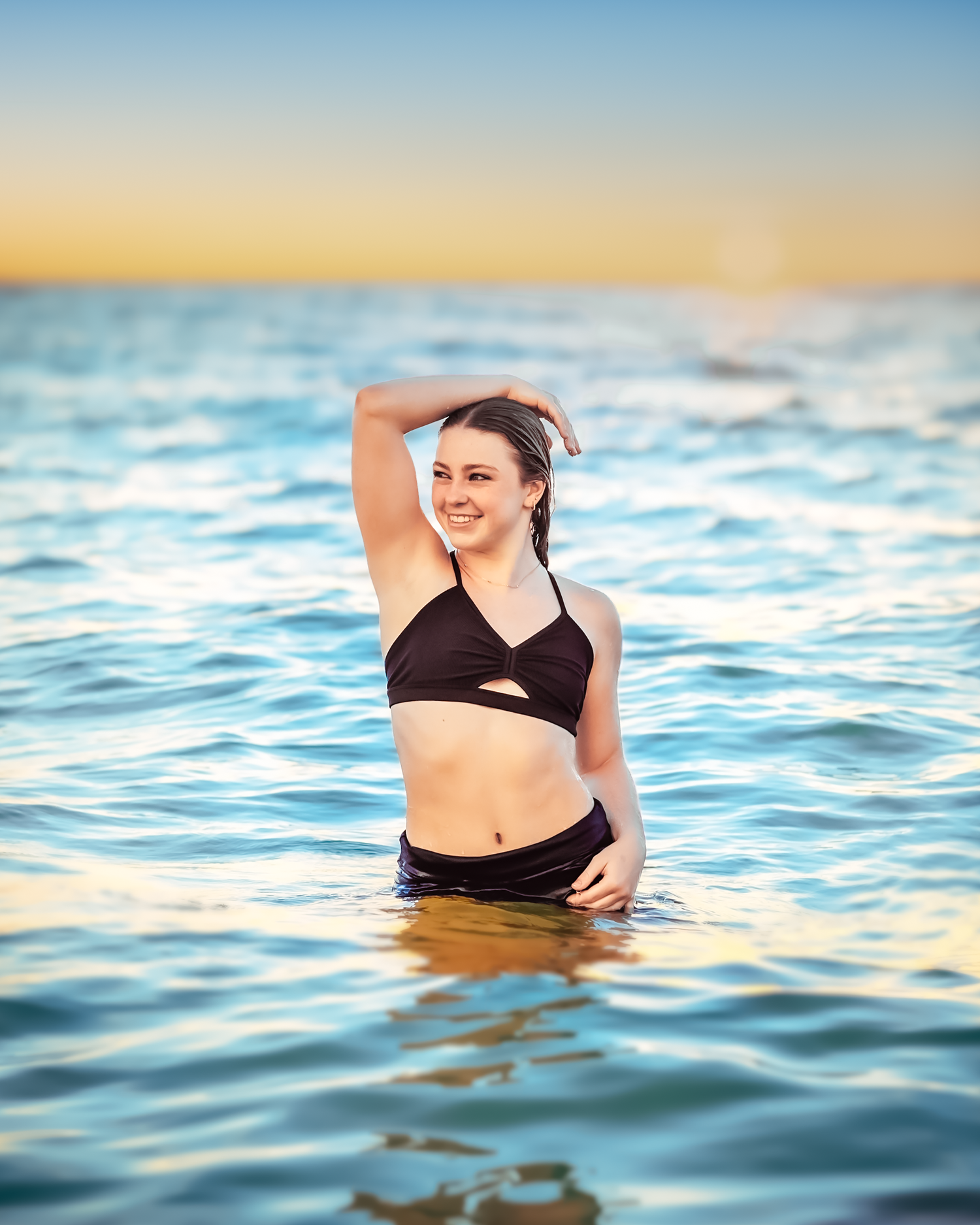 Young woman in black bikini smiling while standing in calm ocean water at sunset.