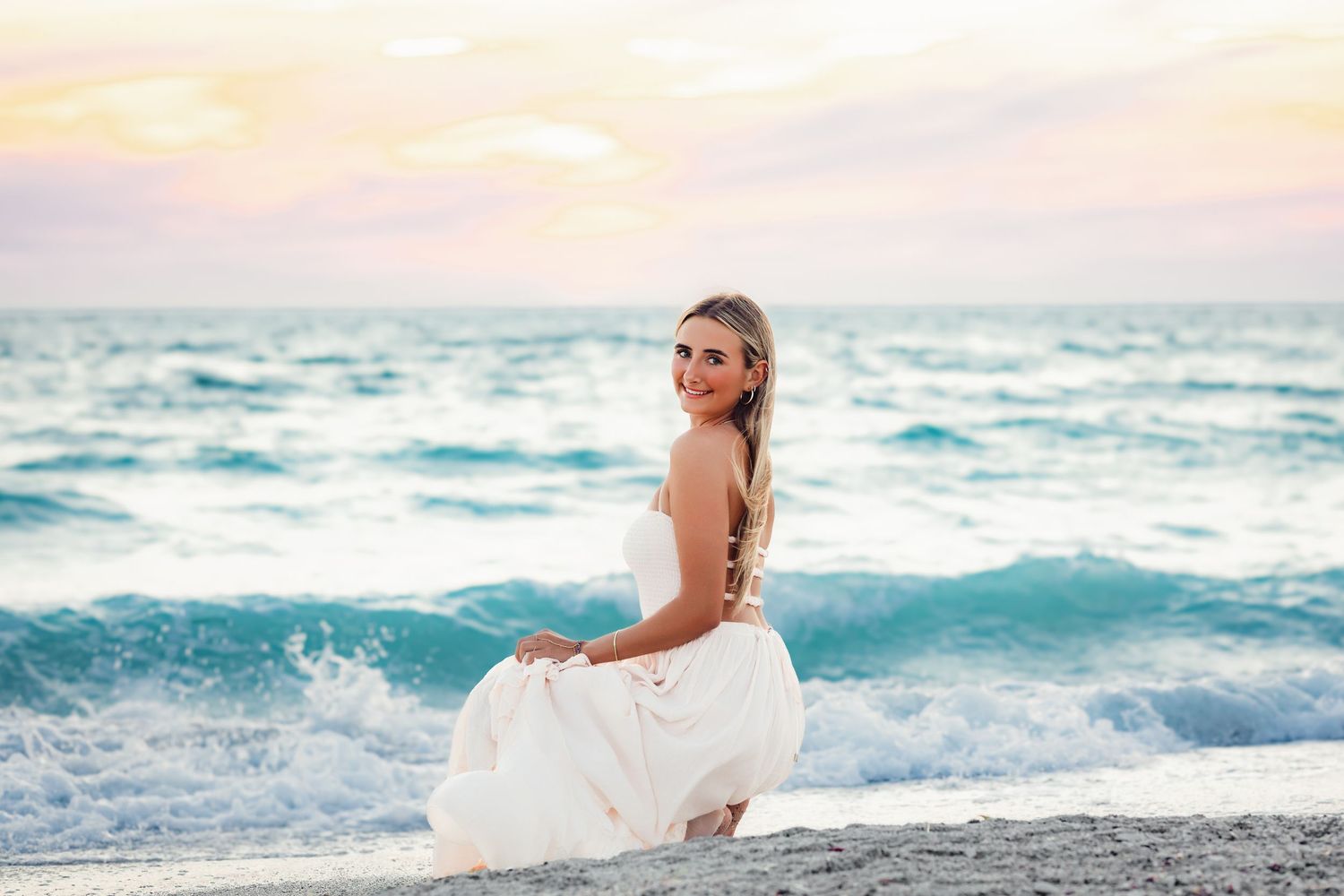 Blonde woman in white dress sitting on beach shore with waves and pastel sunset behind.