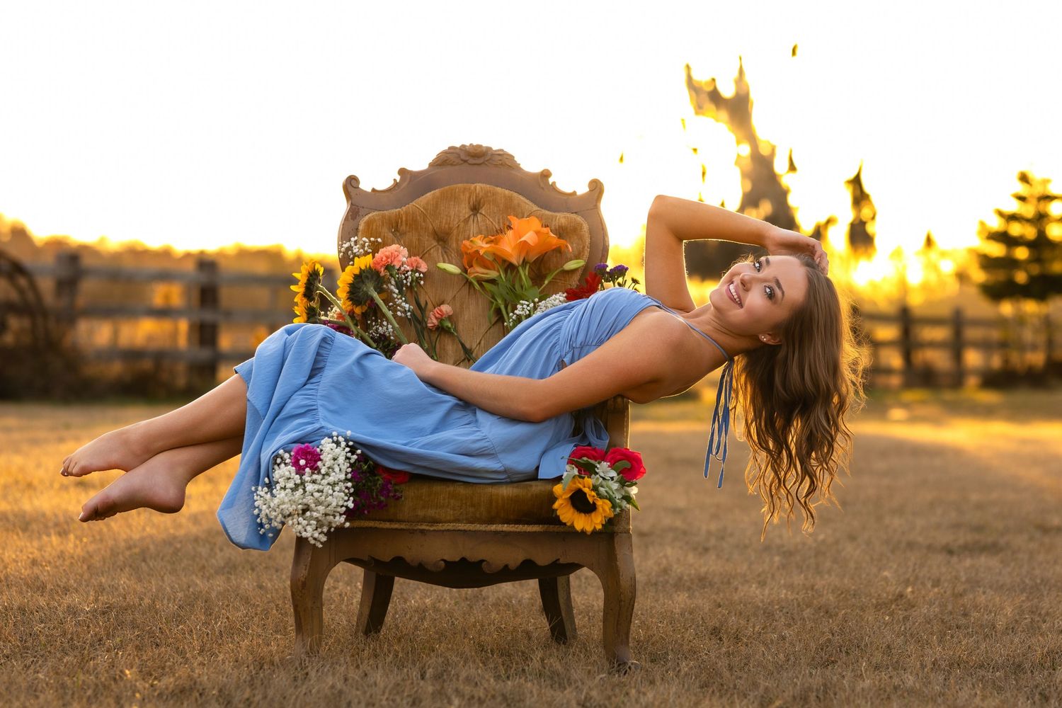 Young woman in blue dress lounging in ornate chair surrounded by flowers at golden sunset.