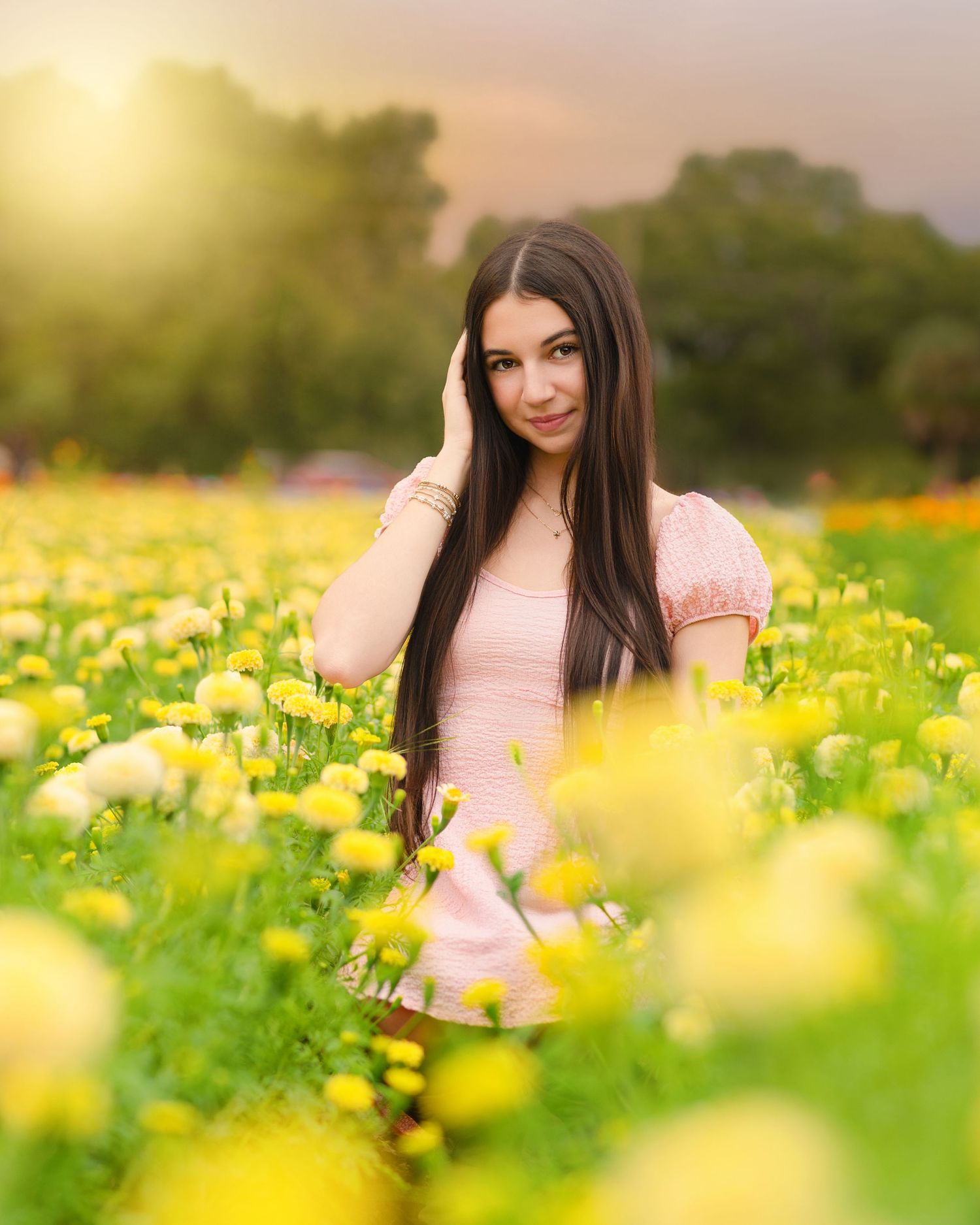 Brunette woman in pink dress sitting in vibrant yellow flower field at golden hour.