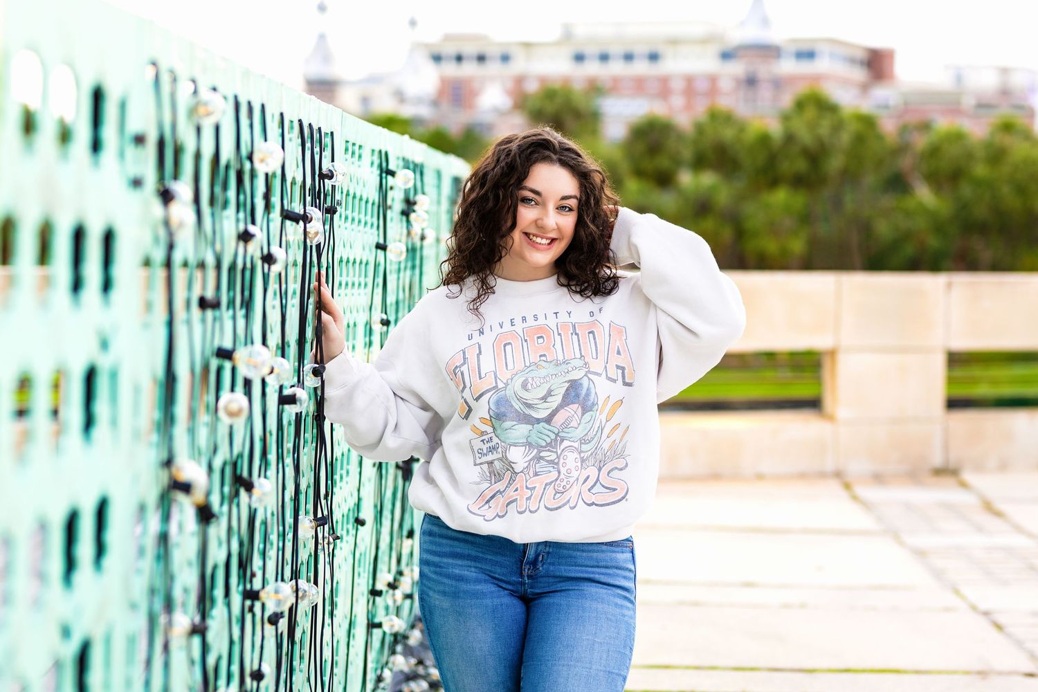 Curly-haired teen girl in Alabama sweatshirt and jeans poses by teal lockers outdoors.