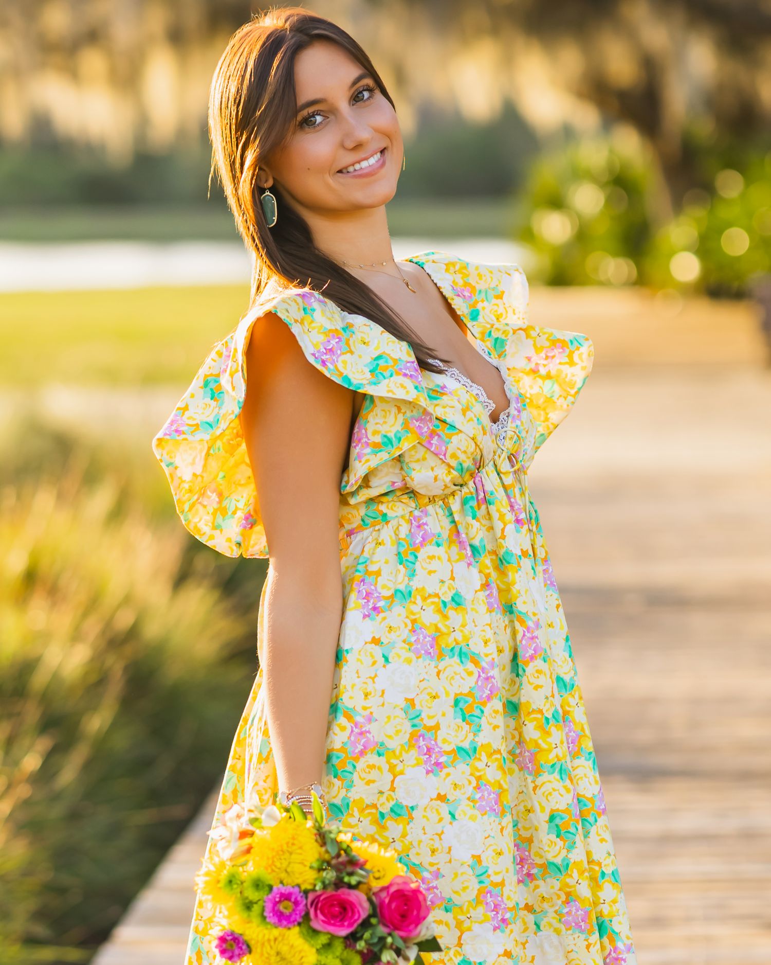 Smiling woman in yellow floral ruffle dress holding colorful bouquet outdoors at golden hour.