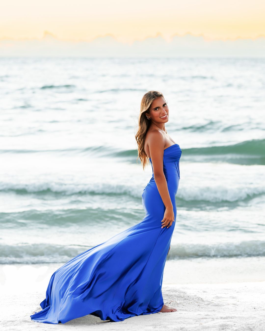 Young woman in flowing royal blue strapless gown posing on sandy beach at sunset.